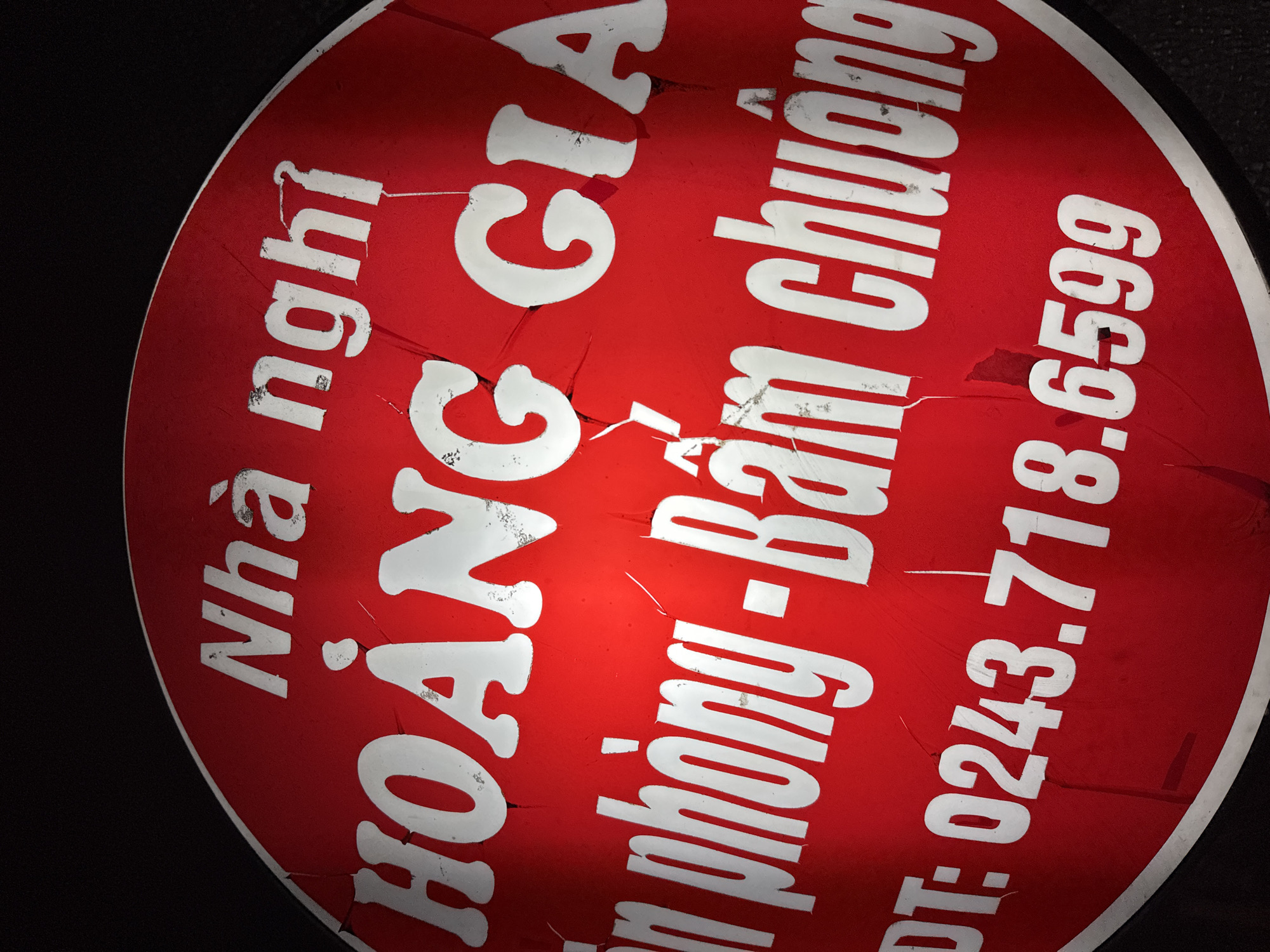 A circular, red sign with white lettering is visible against a dark background. The sign appears to be made of metal or a similar rigid material and has visible cracks and wear marks, suggesting it has been exposed to the elements.
The text on the sign is in Vietnamese. The top line reads "Nhà nghỉ," which translates to "Guesthouse" or "Inn." Below this, in larger, bold lettering, is "HOÀNG GIA," meaning "Imperial" or "Royal."
The next line of text, also in white, is "in phòng - Bám chuong." This appears to be a partial phrase. "in phòng" can mean "rent room" or "in room," and "Bám chuong" might be a phonetic spelling or a specific term related to accommodation.
The bottom line of the sign displays a phone number: "ĐT: 0243.718.6599". "ĐT" is an abbreviation for "Điện thoại," meaning "telephone." The number is a typical Vietnamese landline number for the Hanoi area (indicated by the area code 024).
The overall impression is of a weathered sign for a guesthouse or inn named "Hoàng Gia," located in Hanoi, Vietnam. The sign is likely mounted on a building or at the entrance to the establishment.