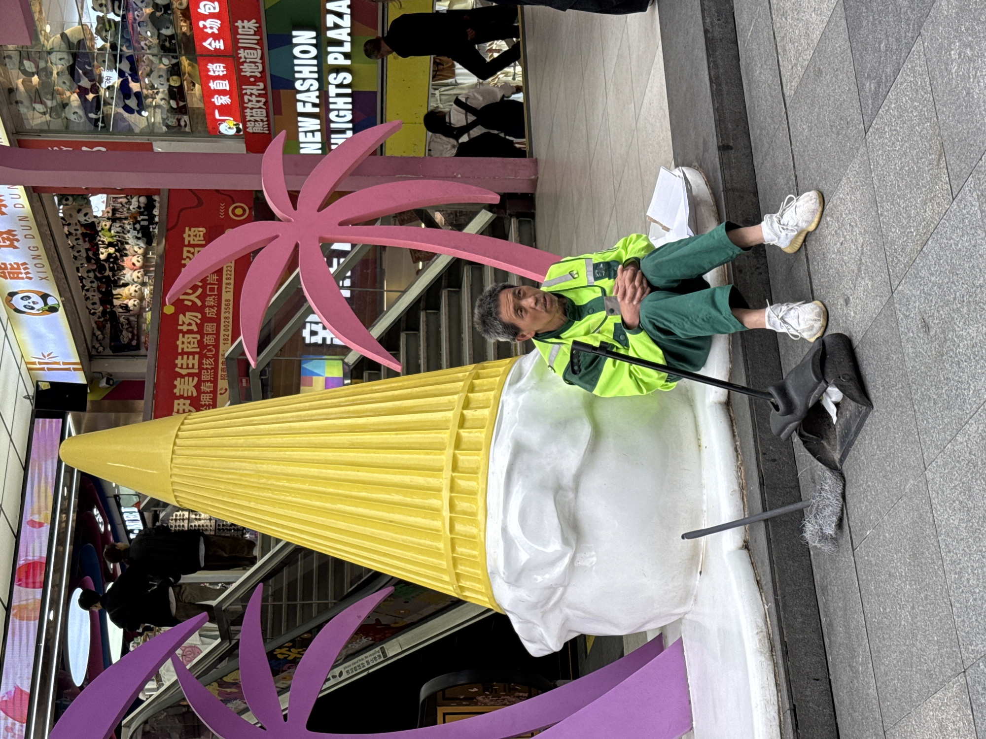 A man wearing a neon green work vest and matching pants is sitting on the edge of an oversized ice cream cone statue in Jinjiang District, China. He has a broom and dustpan next to him. In the background, an escalator leads to a shopping area with signs in Chinese, one of which advertises “NEW FASHION LIGHTS PLAZA”. A sign on the left says "XIONG DUN DUN". Other people are walking in the background.