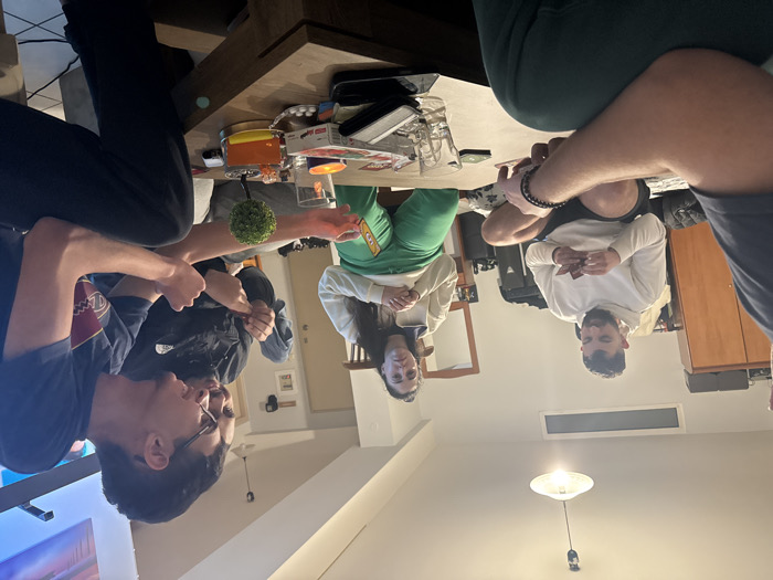 The image depicts five individuals in an indoor residential setting in Plataniás, Greece, engaged in a card game around a rectangular wooden coffee table.
The environment features light-colored walls, an overhead ceiling light fixture with a white shade, a high horizontal window, a large wooden wardrobe on the left, and a mirror over a dresser on the right side of the background. A television screen is partially visible on the far right.
The individuals are:
1. A male subject with his back to the camera in the immediate foreground, wearing a dark short-sleeved shirt and dark pants, with a dark beaded bracelet on his left wrist.
2. A male subject seated on the floor to the left, cross-legged, wearing a light-colored hooded sweatshirt and dark shorts, holding and examining a hand of cards.
3. A female subject seated in the background-center, wearing a light-colored zippered top and green pants, with her hands resting clasped.
4. A male subject to the right-background, partially obscured, wearing a dark jacket with a white logo, holding a small card or object.
5. A male subject with glasses in the right-foreground, wearing a dark t-shirt with a white graphic, extending his left hand to display a green and orange card, while looking towards the center of the table.
The coffee table contains multiple objects: several clear drinking glasses, a small artificial green topiary plant, a lit candle in a metallic holder, an orange container, a dark-cased smartphone, small white circular objects on a wrapper, and various card game components.