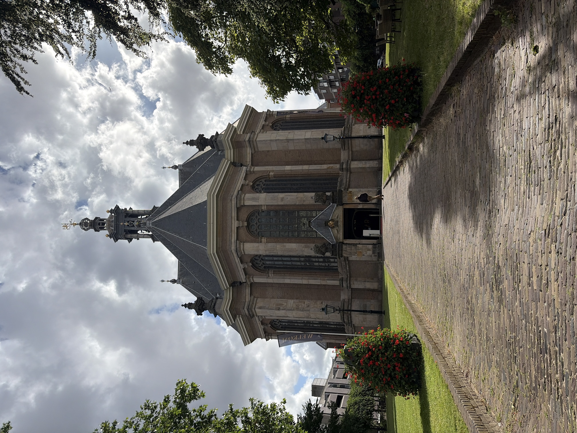 The image shows the Westerkerk church in The Hague, Netherlands. The church is a brick building with a tall, octagonal tower topped with a spire and a weather vane. The facade features arched windows and decorative stonework. A paved walkway leads up to the entrance of the church, flanked by green lawns and bushes with red flowers. A single person is walking away from the camera on the walkway. The sky is partly cloudy. In the background, there are trees and modern buildings.