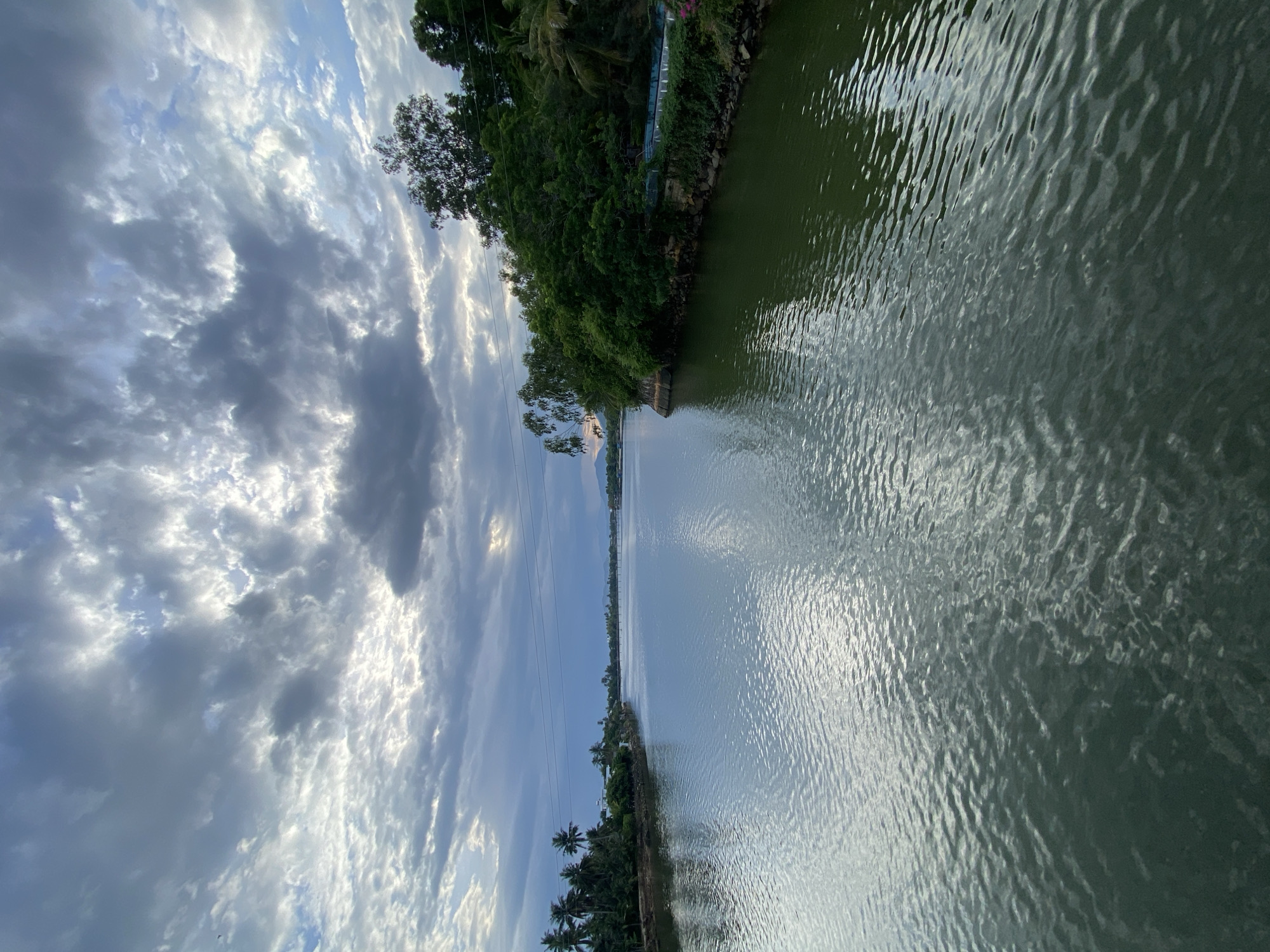 The image depicts a wide river in Nha Trang, Vietnam, under a cloudy sky. The river's surface is calm with ripples reflecting the light. Lush green trees line the right bank of the river, with some houses and more trees visible on the left bank in the distance. A bridge can be seen spanning the river further down. The overall scene is one of natural beauty and tranquility.