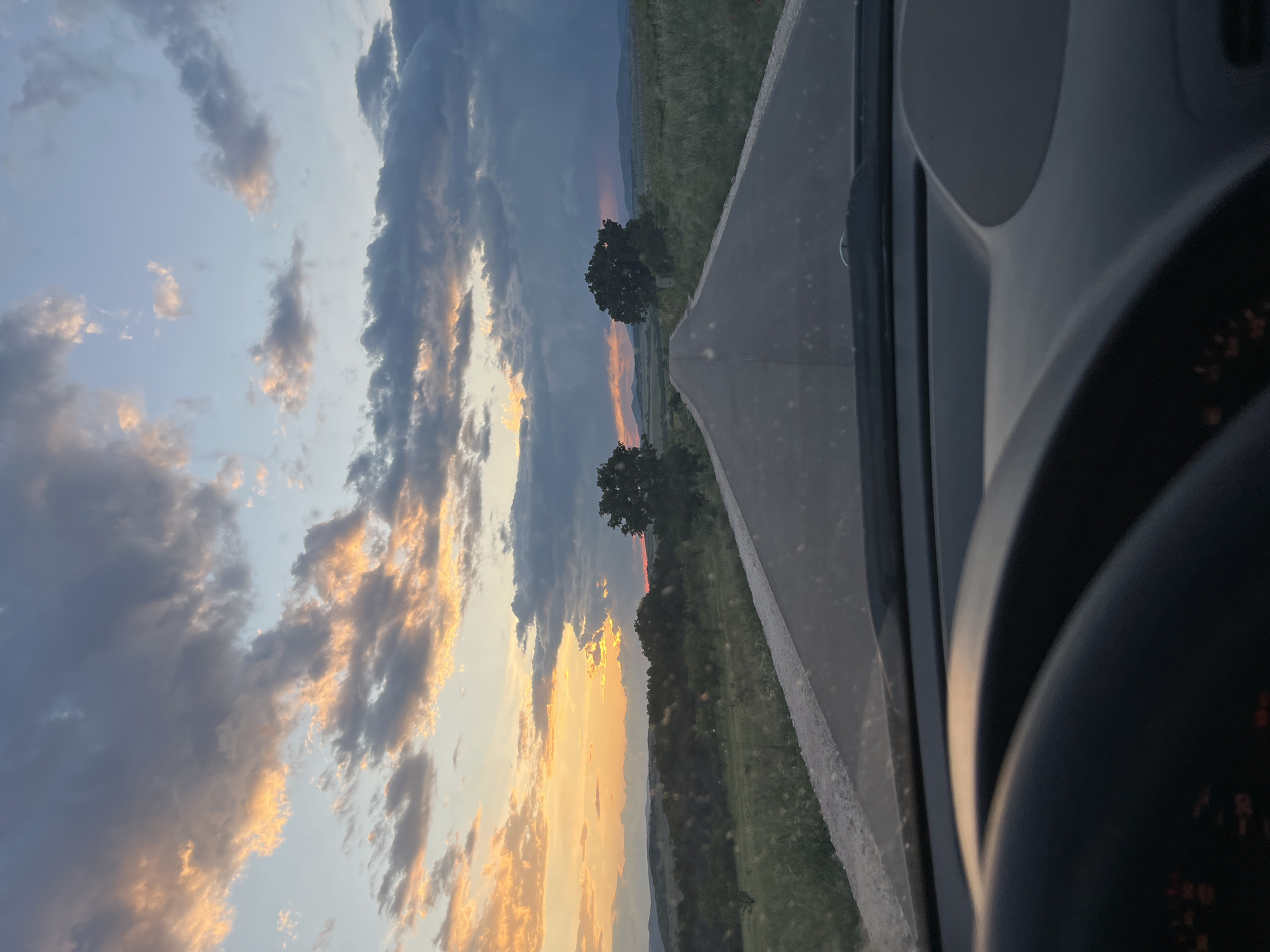 The image shows a view from inside a car on a road, likely in transit. A gray road stretches out, bordered by green fields. Two prominent trees stand near the road. The sky is filled with clouds illuminated by the setting sun, displaying a range of colors from orange to gray. Parts of the car interior are visible, including the dashboard and steering wheel. The image is taken from the city of Smilets, Bulgaria.