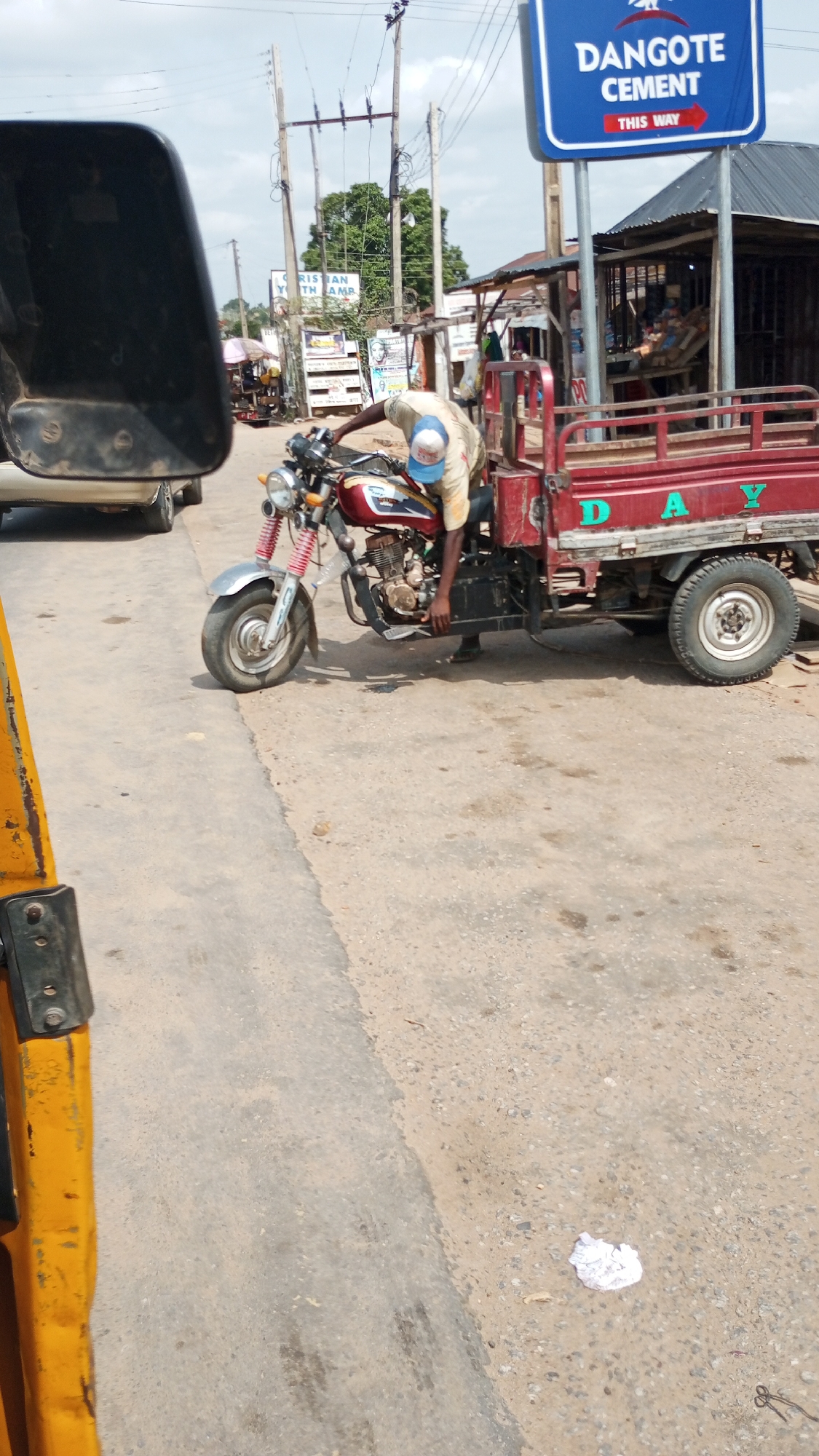 A man in Zango-ndaji, Nigeria, wearing a blue and white baseball cap and a light-colored shirt, is bent over and appears to be working on the engine of a red and silver motorcycle attached to a red three-wheeled cargo carrier. The carrier has the letters "DAY" painted on its side. A blue sign reading "DANGOTE CEMENT THIS WAY" is visible in the background. The scene takes place on a paved road with some debris and a piece of white paper on the side. Power lines and other buildings are visible in the background.