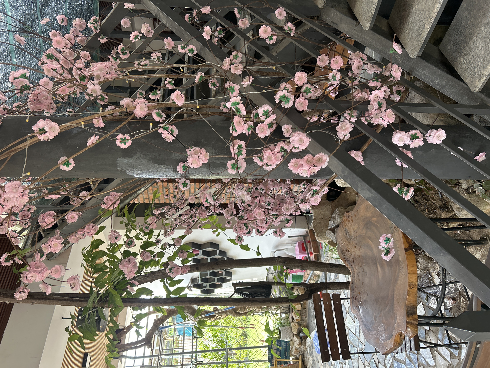 The image shows an outdoor seating area with a wooden table and chairs, decorated with artificial pink cherry blossoms. A staircase with metal railings is visible in the background, with more cherry blossoms attached to it. The area appears to be part of a cafe or restaurant in Nam Nha Trang Ward, Vietnam, with tropical plants and stone landscaping visible. The scene is brightly lit, suggesting it is daytime.