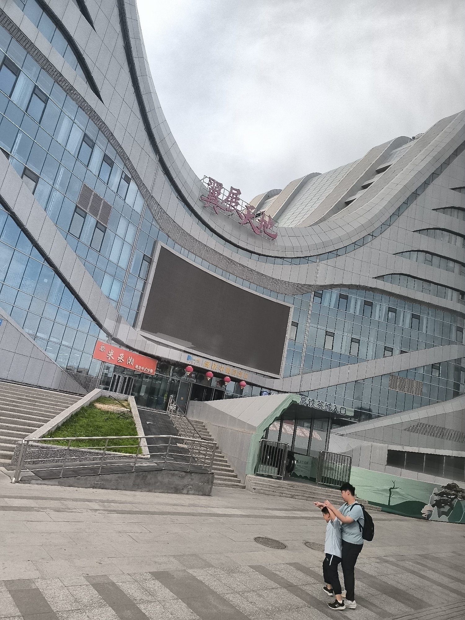 The image depicts a wide-angle view of a modern, architecturally distinctive building in Urumqi, China, likely a commercial or transportation hub, with a man and a child in the foreground. The building's facade is characterized by sweeping curves, a combination of glass windows reflecting a cloudy sky, and grey paneling. A large, dark electronic screen is mounted prominently on the building's exterior.
Above the entrance, a red banner displays Chinese characters. Above that, large pink Chinese characters are affixed to the building's structure. To the right of the main entrance, a smaller, green-roofed structure with glass doors is visible, with Chinese characters above it indicating "High-speed Rail Entrance." Red lanterns hang near this entrance.
In the lower right quadrant of the image, a man and a young child are the primary human subjects. The man, wearing a light blue t-shirt, dark pants, and a backpack, has his arm around the child and is pointing with his other hand towards the left of the frame. The child, dressed in a light grey t-shirt and dark pants, is looking in the direction the man is pointing. They appear to be interacting, possibly navigating or observing something.
The foreground consists of a paved plaza with distinct patterns of light and shadow. A set of wide concrete stairs leads up to a ramp with a metal handrail, bordered by a small patch of green grass. The overall environment suggests an urban setting on an overcast day.