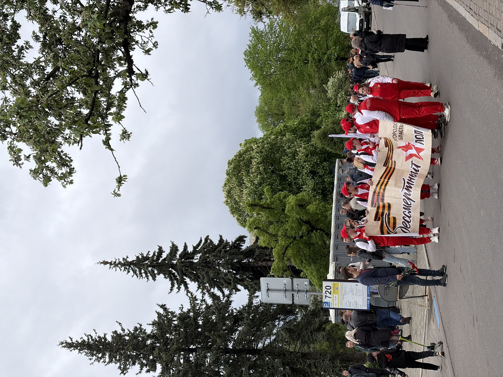 The image shows a group of people walking down a city street, carrying a banner with a ribbon and Cyrillic script. The people are wearing red and white tracksuits. On the side of the road, people are watching. There is a bus stop sign with the number 720. Trees are on both sides of the street. The sky is overcast.