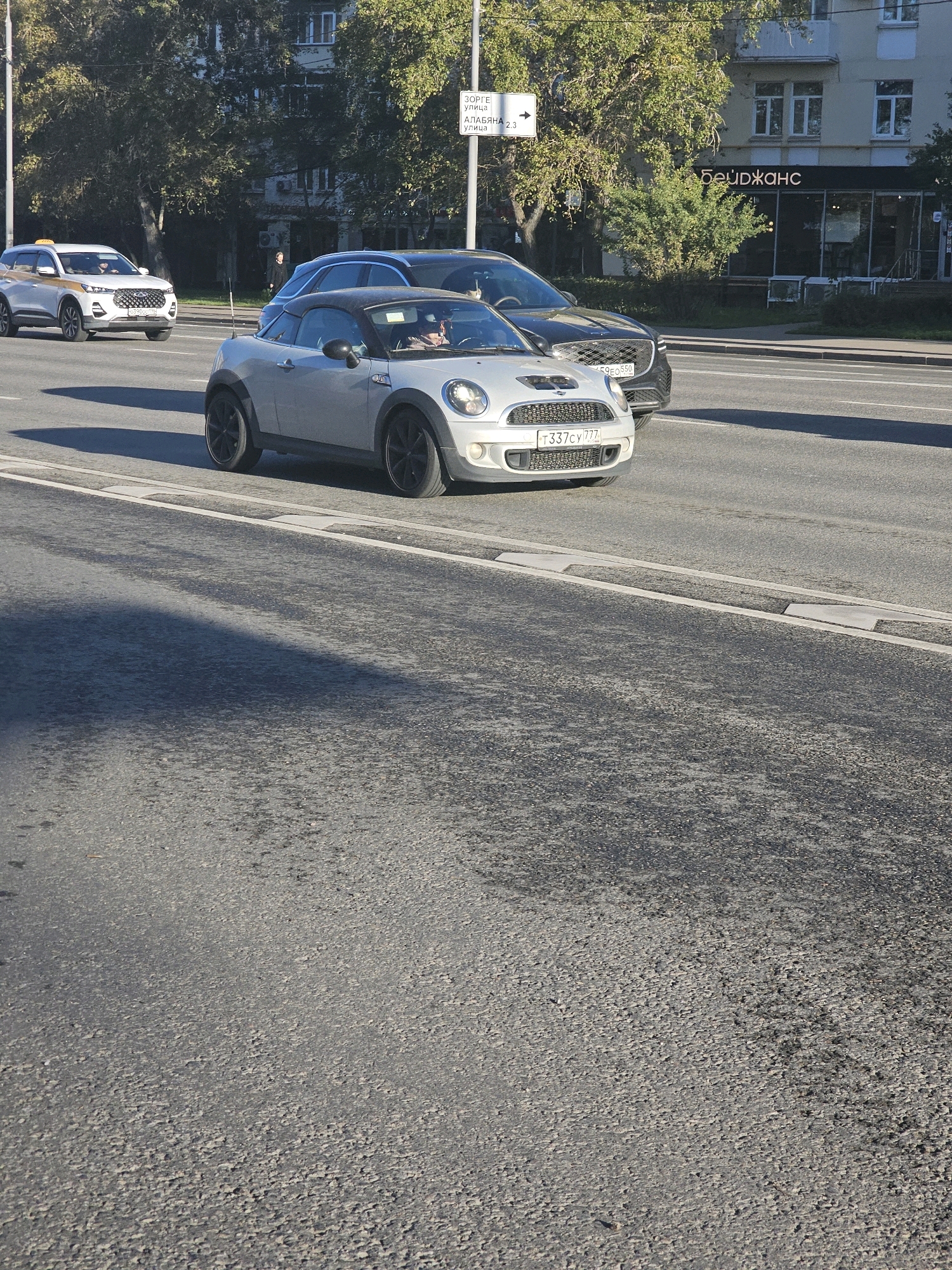 The image captures a street scene in Moscow, Russia, on a clear day. In the foreground, a silver Mini Cooper convertible is driving on a paved road. The car's license plate is visible and reads "T337CY 777". Behind the Mini Cooper, a dark-colored SUV is also on the road. To the left of the Mini Cooper, a white SUV, possibly a taxi, is visible. In the background, there are trees, a building with windows, and a street sign. The street sign has Cyrillic text and an arrow pointing to the right, indicating directions to "ЗОРГЕ УЛИЦА" (Zorge Street) and "АЛАБЯНА 2,3" (Alabyan 2,3). The building in the background has a sign with Cyrillic text that appears to read "БЕЙДЖАНС" (Beidzhan's). The overall scene depicts urban traffic and architecture.