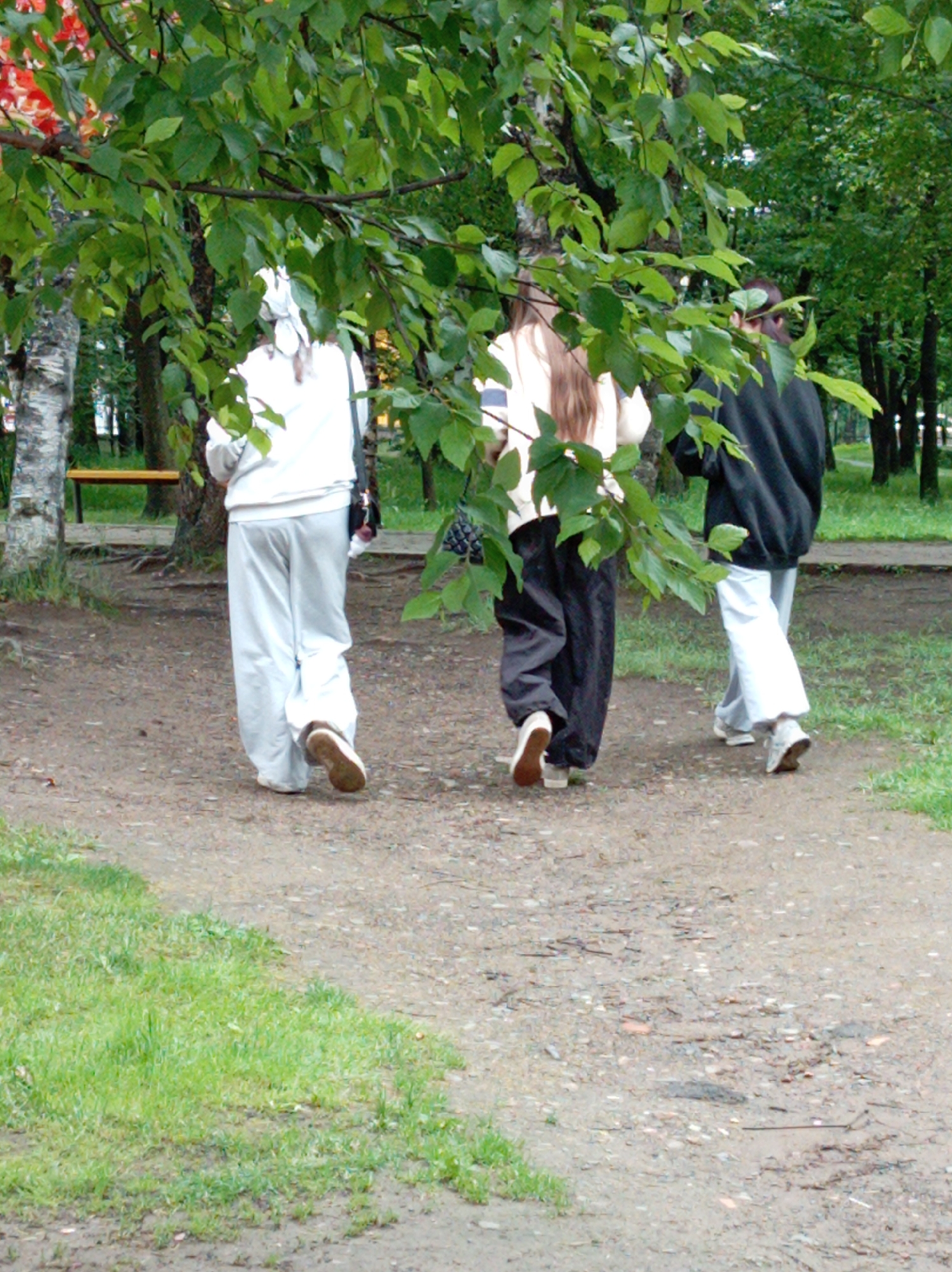 Three individuals are walking away from the camera on a dirt path in a park. The person on the left is wearing a white hooded sweatshirt and loose white pants. The middle person is wearing a light-colored top and loose dark pants, with long brown hair. The person on the right is wearing a dark hooded sweatshirt and white pants. All three are wearing casual shoes. The path is surrounded by trees with green leaves and some grassy areas. In the background, there are more trees and some distant structures that appear to be part of the city. The lighting suggests it is daytime, possibly overcast. The scene is likely located in a public park within the city of Artyom, Russia, given the presence of urban elements mixed with natural surroundings.