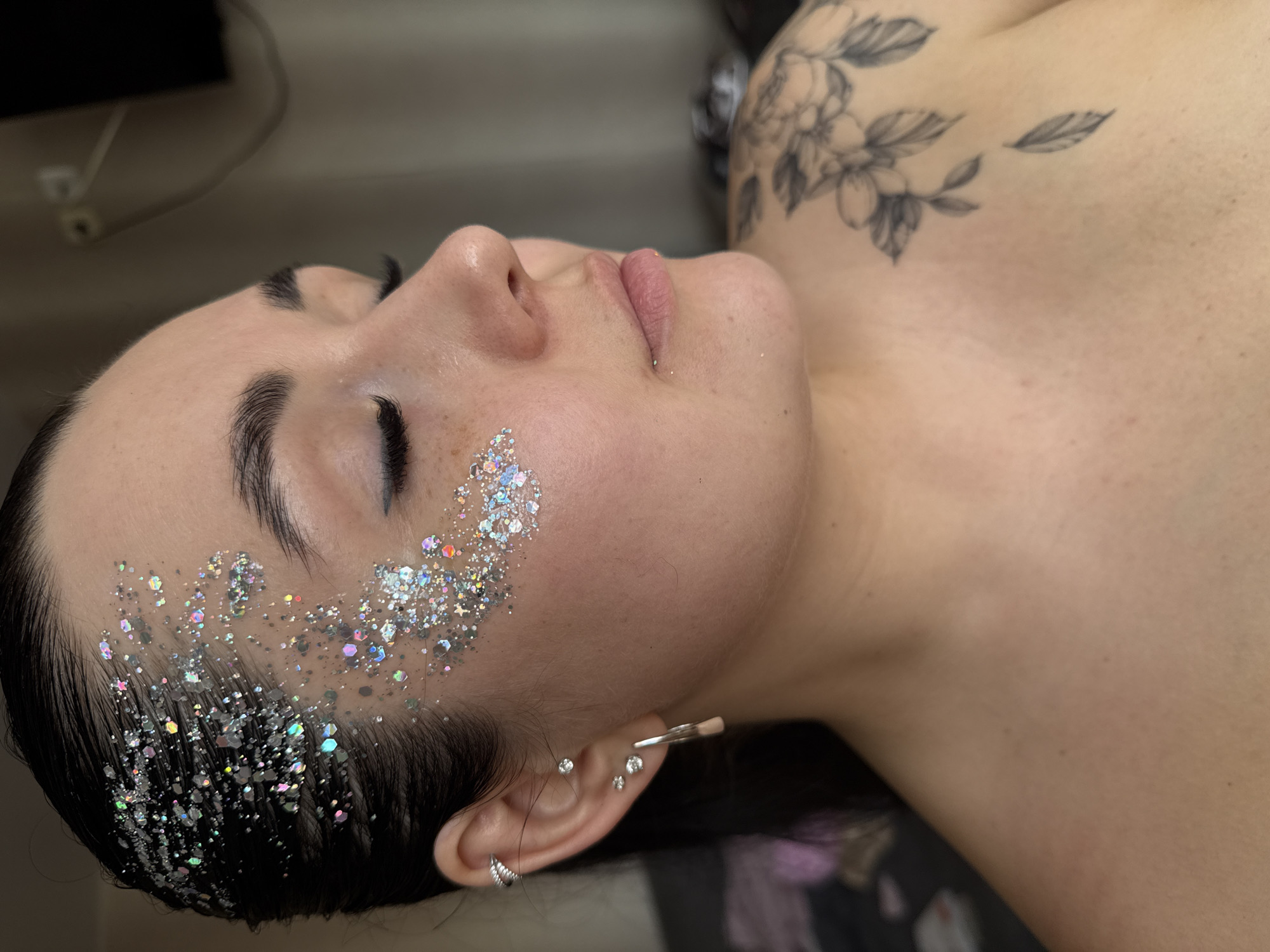 The image presents a close-up, high-angle portrait of a person, likely female, lying down with their eyes closed, suggesting a state of rest or relaxation. The subject's head is tilted slightly to their left, prominently displaying the right side of their face, neck, and shoulder.
A striking application of chunky, iridescent silver glitter covers a significant portion of the left temple, cheekbone, and along the hairline, sparkling brightly under the light. The person has dark hair, dark and well-defined eyebrows, and naturally pink, full lips. Several silver piercings adorn her left ear, including two small stud earrings on the lobe, another stud further up, and a bar or cuff-style piercing on the upper helix.
A detailed black and grey floral tattoo, depicting flowers and leaves, is visible on the person's right shoulder and upper back. The skin appears smooth with a few scattered freckles on the shoulder. The background is mostly dark and out of focus, creating a shallow depth of field that keeps the focus on the subject. No text is visible in the image. The location context for this image is Chaikovsky, Russia.