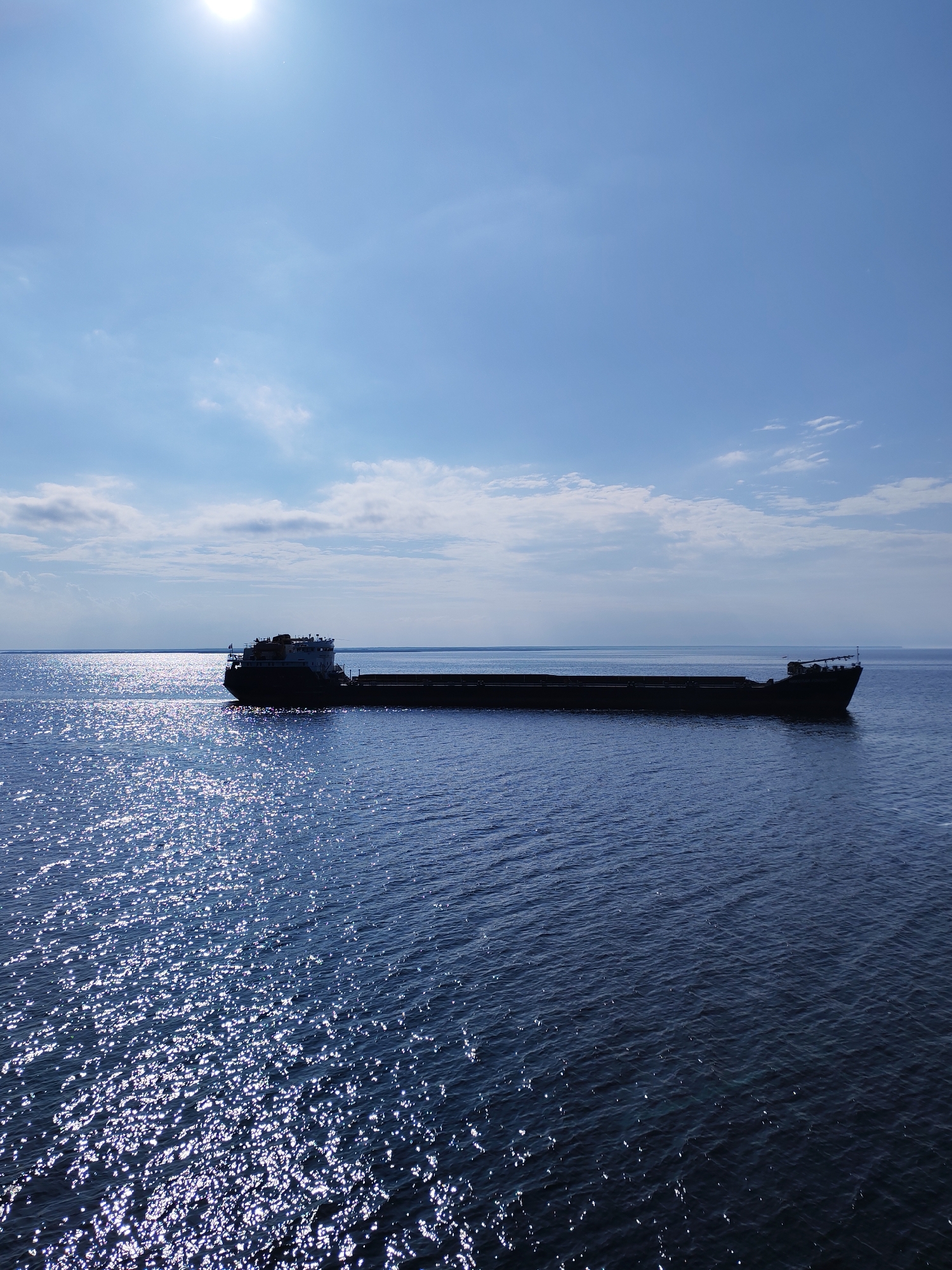 The image depicts a long, dark, empty cargo ship on a body of water, likely a sea or large lake. The water surface is highly reflective, with sunlight creating a shimmering effect. The sky is blue with scattered clouds. The sun is visible as a bright spot at the top of the image. There are no people visible. The location is unknown.
