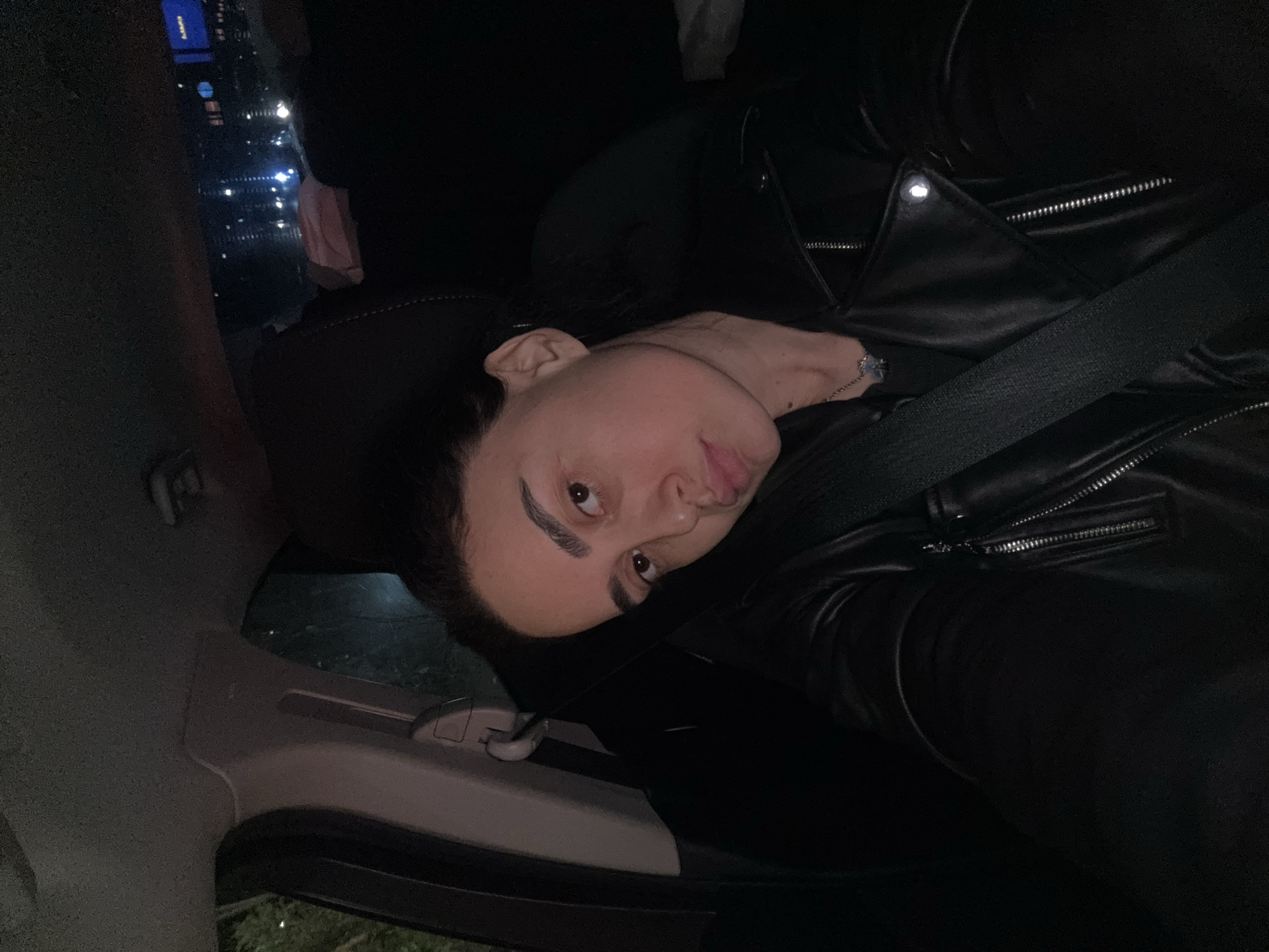A close-up, vertically oriented selfie captures a young adult inside a car at night. The person, positioned in a car seat, looks directly at the camera with a slightly pouty or serious expression. They are wearing a black leather jacket with silver zipper details and a prominent snap button on the lapel. A black seatbelt is fastened across their chest. A light-colored bracelet with a distinctive charm is visible on their wrist.
The car's interior appears dark, with portions of a light-colored door panel and window frame visible. Outside the car window, the scene is dark, indicative of nighttime, with blurred distant city lights and what appears to be a street or building. One small, blue rectangular light in the background might be a screen displaying faint, illegible white text.
The individual has dark hair and distinct, dark eyebrows. The image's lighting suggests it was taken with a flash or in low light conditions within the vehicle, contrasting with the dark background. The provided context indicates the location is Tbilisi, Georgia.