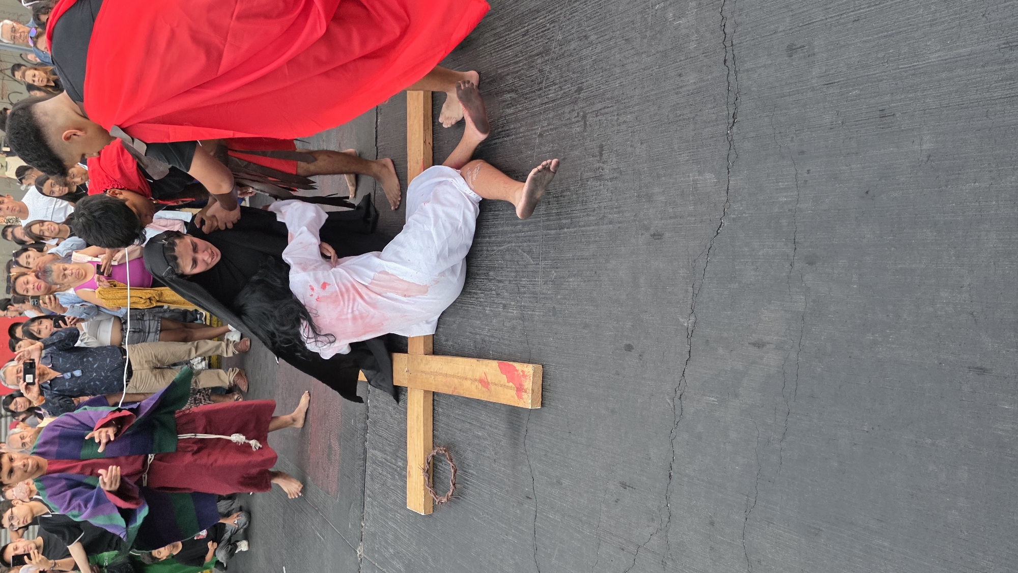 This image depicts a scene of a religious reenactment or performance, likely related to the crucifixion of Jesus Christ. The setting appears to be an outdoor public space with a crowd of onlookers. The central figures include a person portraying Jesus, lying on a wooden cross, and several other individuals dressed in costumes that resemble Roman soldiers or biblical characters. The person on the cross is wearing a white garment with apparent bloodstains. The "soldiers" are in Roman-like attire and appear to be interacting with the person on the cross. The crowd is observing and taking photos or videos with their phones. A crown of thorns is visible near the cross. The location is identified as Unknown, Unknown.