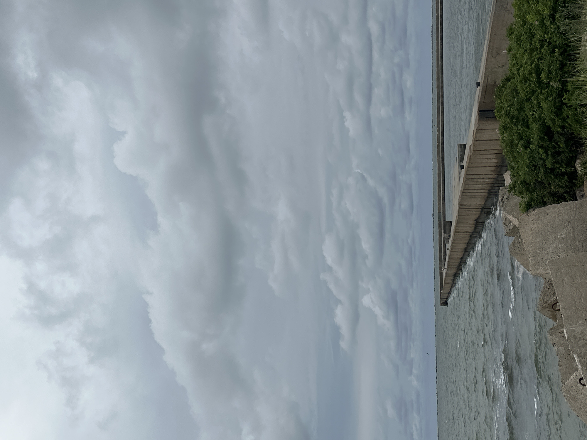 A concrete pier extends into the Baltic Sea under a cloudy sky. The water is choppy with whitecaps. The pier is constructed of large concrete blocks and is visible in the foreground and middle ground, curving to the right. A patch of green vegetation is present on the right side of the foreground, in front of the pier. In the distance, another, parallel concrete structure is visible across the water. The sky is filled with heavy, gray clouds. The scene appears to be at the Liepaja Port in Liepaja, Latvia. There are no people visible in the image.