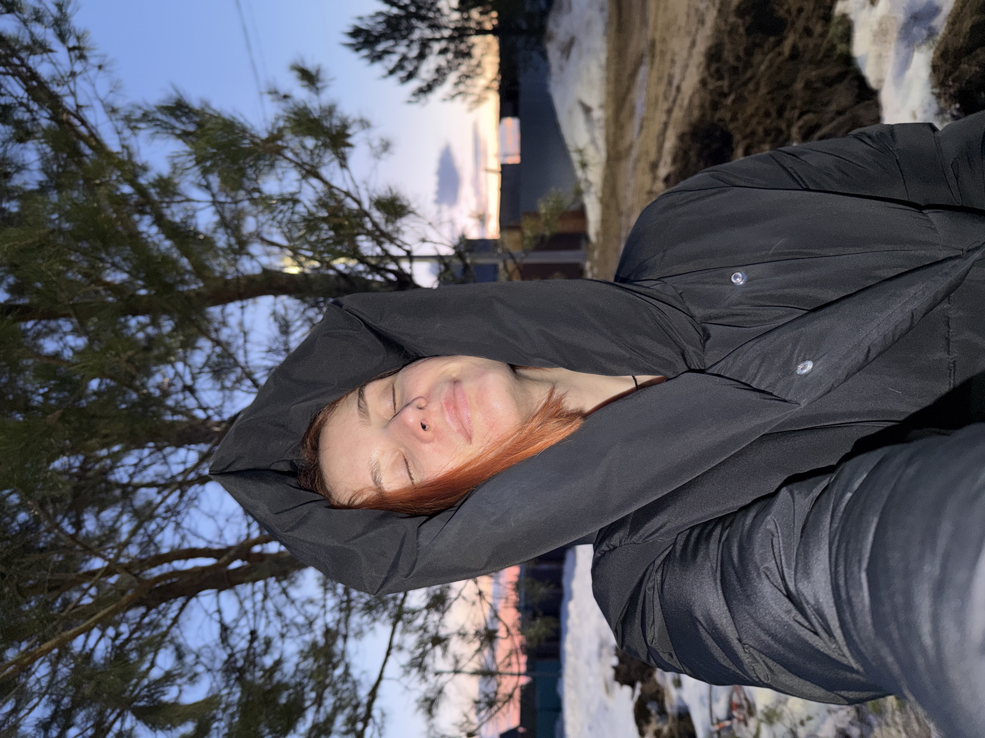 A woman with reddish hair and a fair complexion is lying down outside, wearing a black hooded jacket. Her eyes are closed, and she has a slight smile. She is lying on her back, with trees visible to the left and a snowy area with bare ground to the right and behind her. The background includes a sunset sky. This is a winter or early spring scene. The location appears to be in a residential area of Chaikovsky, Russia, given the presence of snow, trees common to the region, and glimpses of buildings in the background.