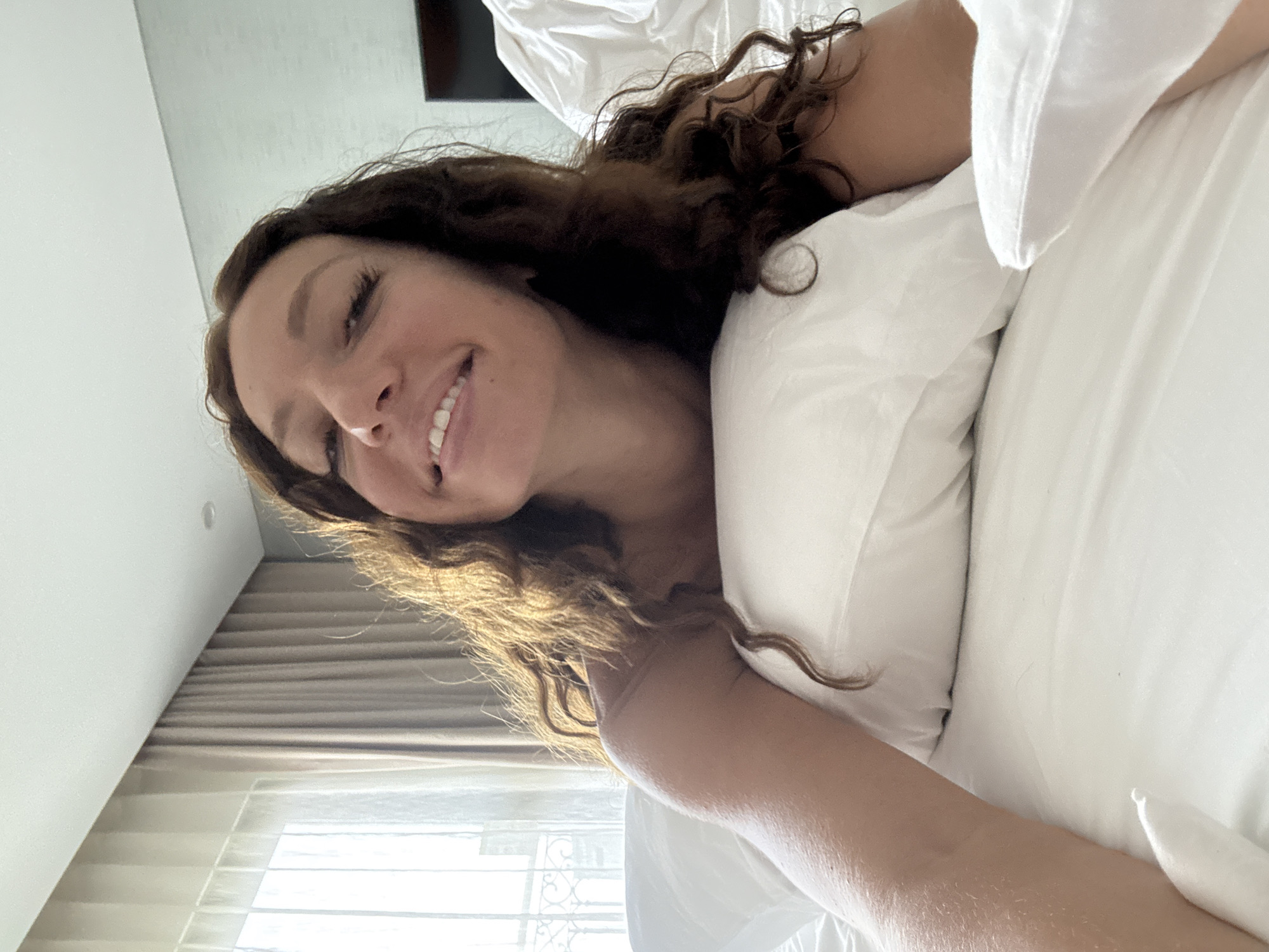 A woman with long, curly brown hair is lying on a white pillow and white sheets. She is smiling. The room has a window with beige curtains and a view of what appears to be city buildings. Based on the prompt, this is Đà Nẵng, Vietnam.