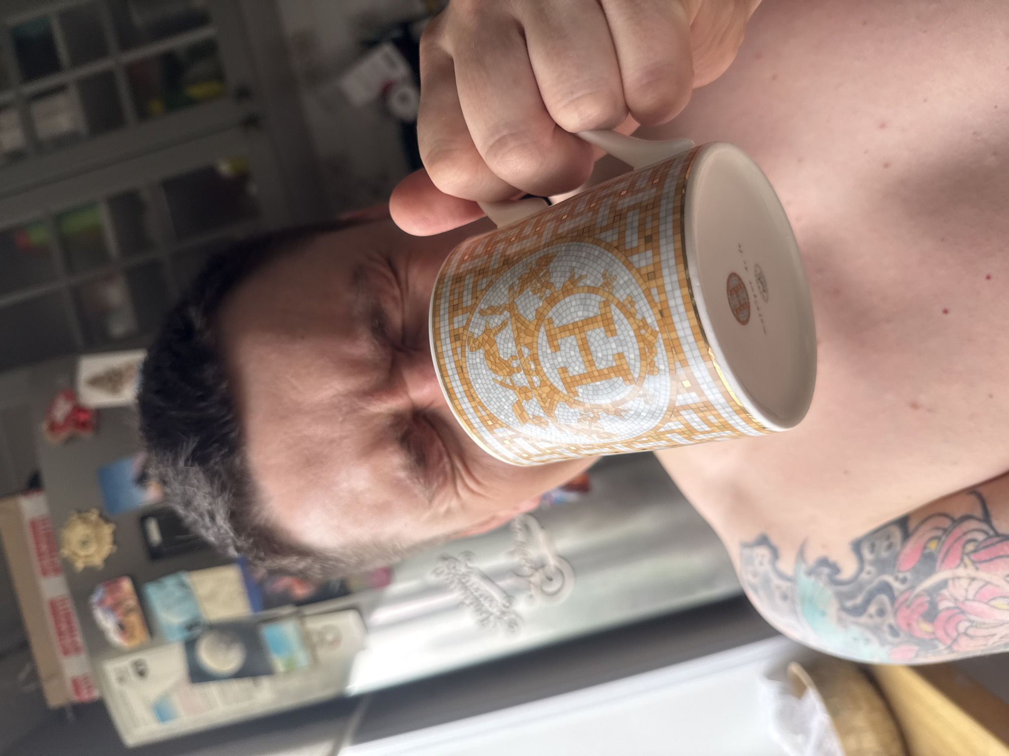 The image is a close-up, Dutch angle, medium shot of a person drinking from a mug in what appears to be a kitchen. The person is shirtless, revealing tattoos on their left arm and shoulder. Their eyes are squinted as they take a sip from the mug, which features a mosaic pattern with an "H" logo. The background shows a refrigerator with magnets and kitchen cabinets. The overall lighting suggests an indoor setting, possibly during the day due to the natural light filtering in from the left. The scene is likely a domestic environment, not specifically identifiable as Vladivostok, Russia, as no distinctive landmarks or cultural elements are visible.