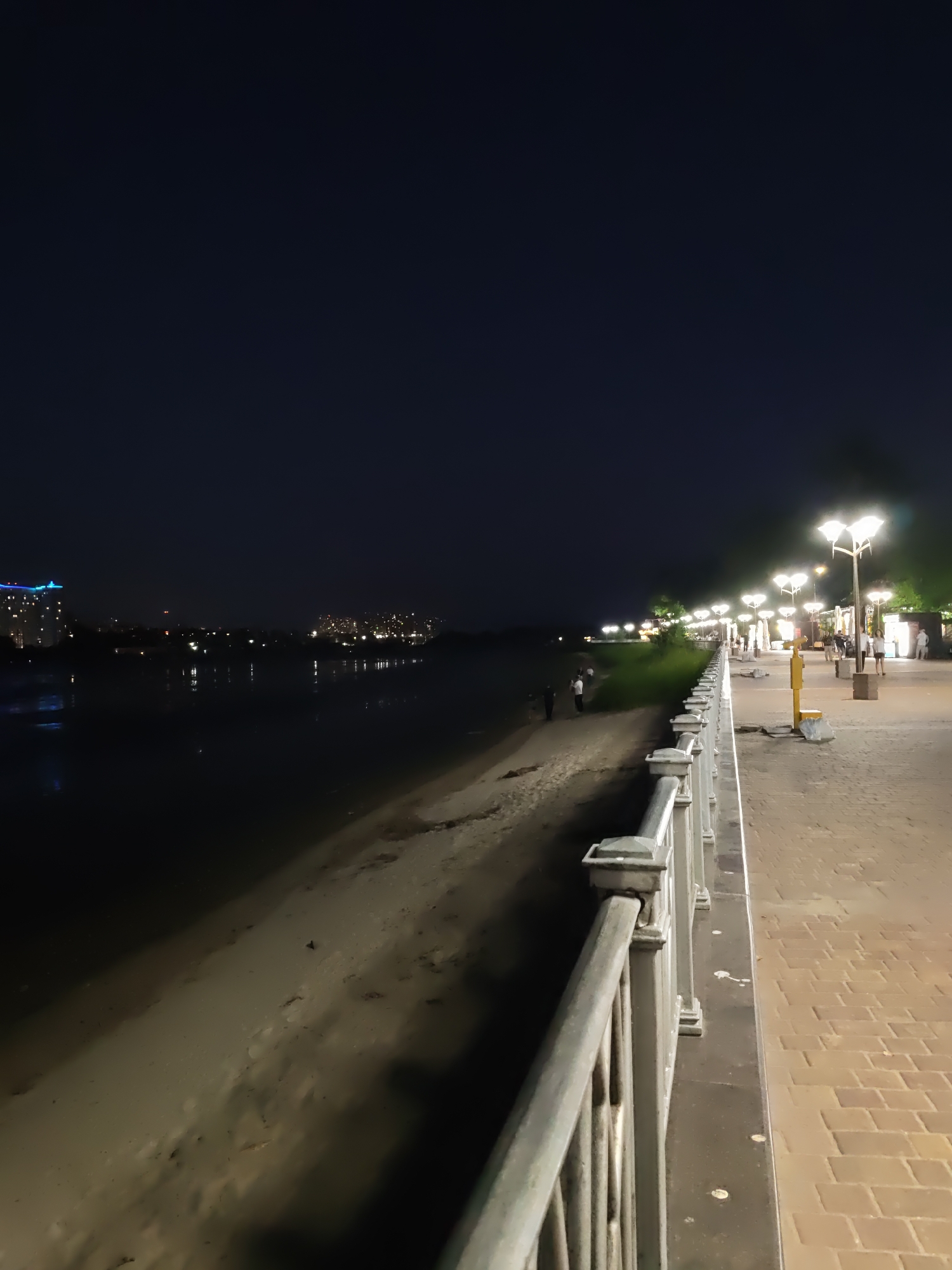 The image depicts a nighttime scene of a riverfront promenade in Rostov-on-Don, Russia. On the right side of the image, a paved walkway with brick-like patterns is illuminated by a row of ornate streetlights. A metal railing separates the walkway from the sandy riverbank on the left. Several people are visible walking along the promenade and on the riverbank. Across the river, the city skyline is illuminated with lights from buildings and reflections on the water. The sky is dark, indicating it is nighttime.