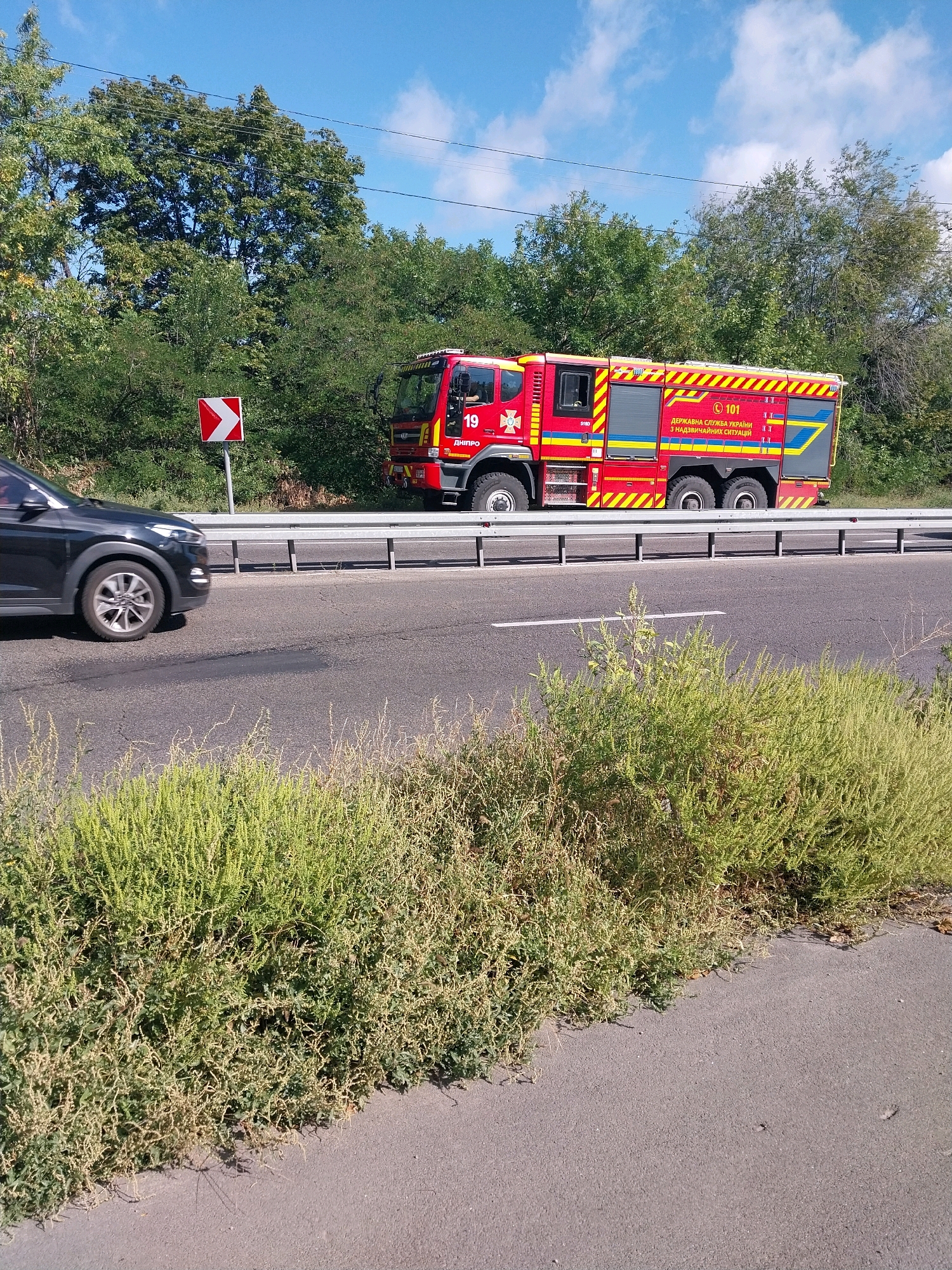 A red fire truck with the number 19 and Ukrainian emergency service markings is driving on a highway in Dnipro, Ukraine. A black SUV is driving in front of the fire truck. The scene is on a sunny day with blue sky and white clouds. The highway is bordered by a metal guardrail and dense green trees. There is also a red and white directional sign to the left of the road.