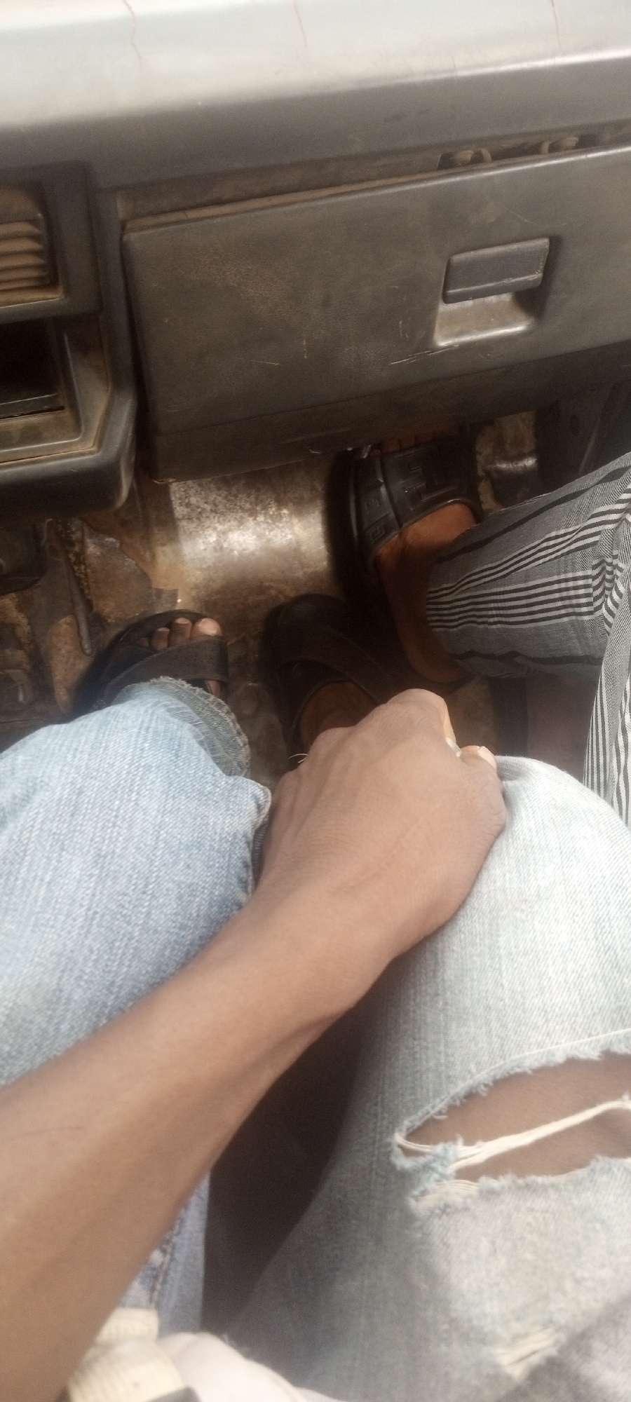 The interior of a vehicle is shown from an elevated perspective. Two pairs of legs are visible, clad in jeans, next to each other. The left leg is in light-wash ripped jeans and a sandal, while the right leg is in dark-colored footwear and striped pants. Underneath the legs is the worn floor of the vehicle. Above, a gray glove box is visible. The location is assumed to be Yelwa, Nigeria, but there is no specific evidence in the image to verify this.