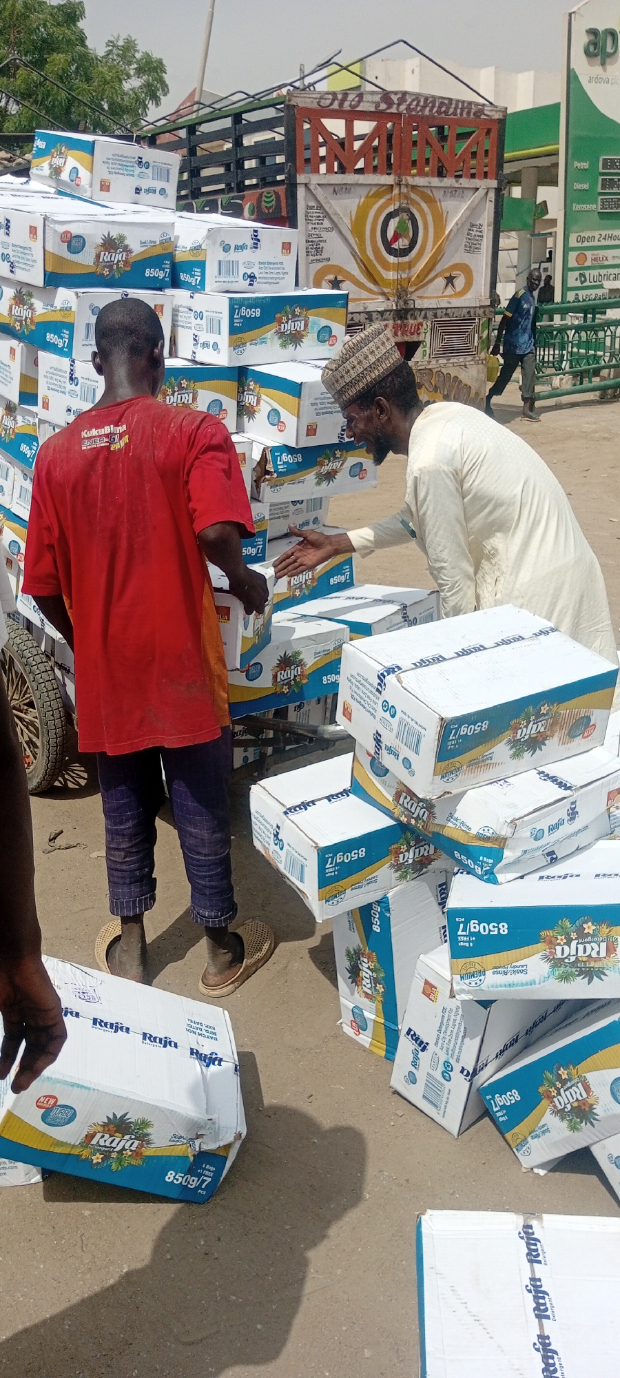 In Sabon Gari West, Nigeria, two men are handling boxes of Raja detergent, each labeled as 850g/7. One man wears a red shirt and dark pants with sandals, while the other wears a light-colored robe and a patterned cap. A truck loaded with more detergent boxes is visible in the background. The boxes are also stacked on a cart, and scattered on the ground. An Ardoav petrol station is also visible. A third man is walking behind a fence.