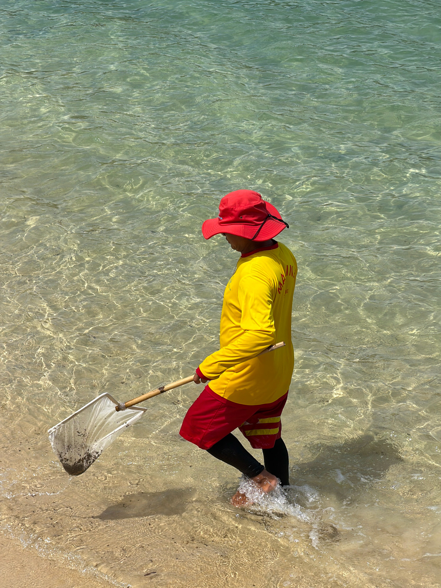 A person wearing a red hat and a long-sleeved yellow shirt with red trim and red shorts is walking in shallow, clear water at the edge of a sandy beach. The person is holding a net with a wooden handle, which contains dark debris. The person's legs are submerged in the water, creating splashes. The scene appears to be a public beach, possibly in Nha Trang, Vietnam, given the clear turquoise water and sandy shoreline. The sun is shining, creating dappled light on the water's surface.