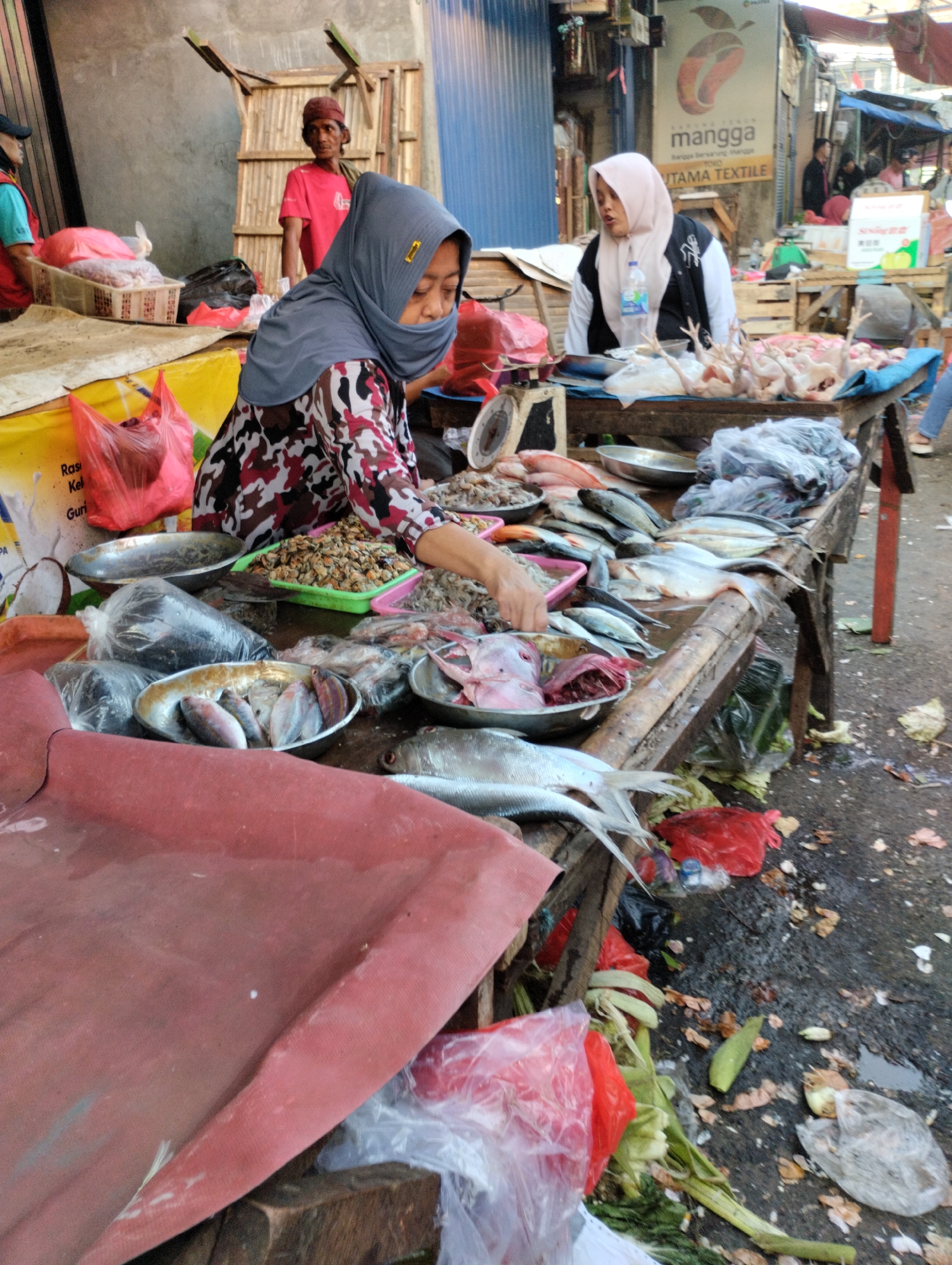 The image captures a bustling scene at a market stall, likely in Bekasi, Indonesia. A woman wearing a grey hijab and a patterned shirt is shown tending to a selection of fresh fish and seafood. Her hands are visible as she appears to be arranging or inspecting the produce on a wooden table. The stall is laden with various items for sale, including different types of fish, shellfish in a green container, and bags of other goods. In the background, other market-goers are visible, including a man in a red shirt and another woman in a light-colored hijab. The environment appears to be an open-air market with stalls made of wood and corrugated metal. The ground is wet and littered with debris, indicating typical market conditions. The overall impression is one of a busy and active marketplace.