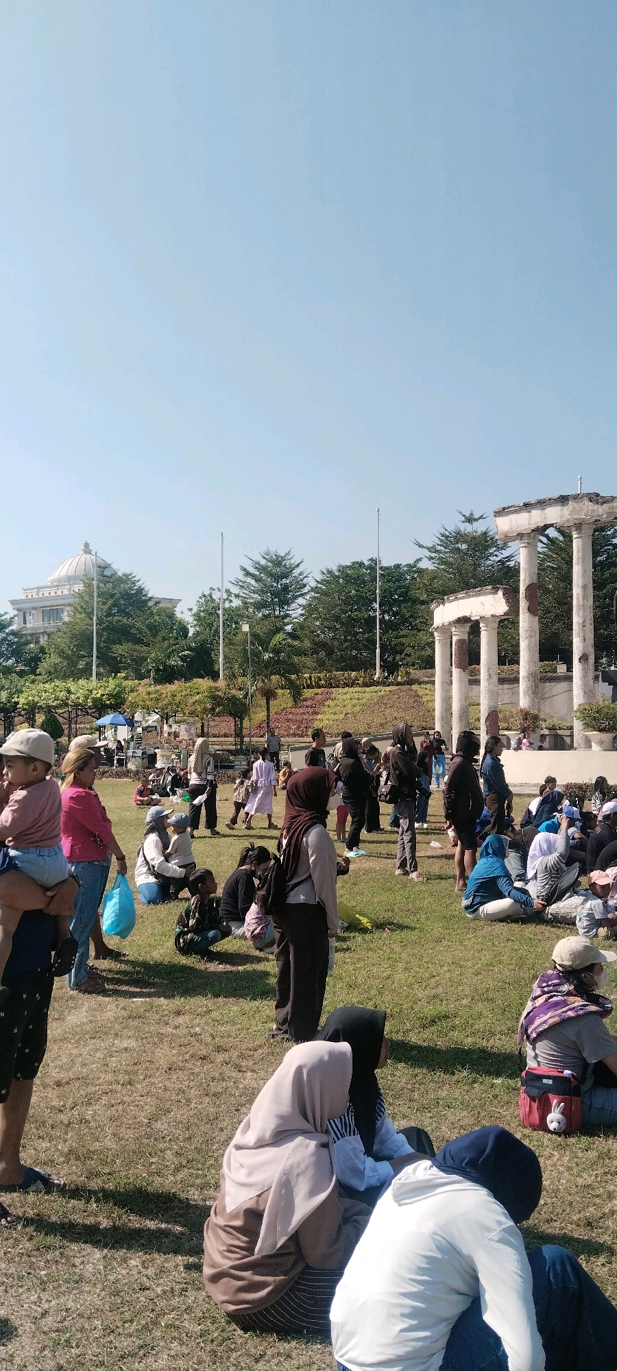 A group of people are gathered on a grassy lawn under a clear blue sky, in what appears to be a park area with some classical-style ruins in the background. A large building with a dome is visible in the distance. The people are diverse in age and are dressed casually, many wearing hijabs, suggesting a Muslim-majority population. Some are standing, while others are sitting or crouching on the grass, some with children.
The ruins consist of several large, broken white pillars and fragments of walls, hinting at a historical or memorial site. Trees and landscaping are visible around the ruins and the distant building, with some areas of colorful flowers. The overall impression is a public gathering or event taking place outdoors on a sunny day.
The location is identified as the City of Surabaya, Indonesia. This specific area, with the ruins and the dome-shaped building in the background, is characteristic of Surabaya's Tugu Pahlawan (Heroes Monument) complex, a significant historical and recreational site. The gathering on the lawn could be for leisure, a public event, or a religious observance. The presence of food vendors in the background suggests it might be a public space that also accommodates small businesses catering to visitors.