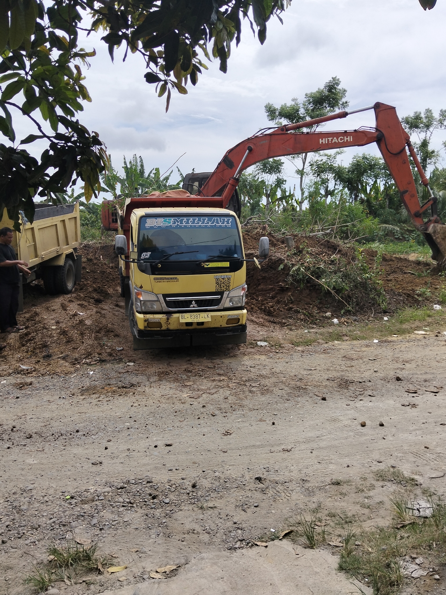 In the city of Payagaboh, Indonesia, a construction site is depicted with an excavator and two dump trucks. The excavator, identified by the "HITACHI" logo, is orange and actively engaged in moving dirt and debris. A yellow dump truck is positioned in front of a pile of excavated earth, appearing to be receiving or having just received material. To the left, another yellow dump truck is visible, partially obscured by foliage. A man, dressed in dark clothing and sandals, stands near the dump truck on the left, gesturing with his hands. The environment consists of a dirt road, piles of earth, scattered branches, and lush green vegetation, including banana trees, in the background under a cloudy sky.