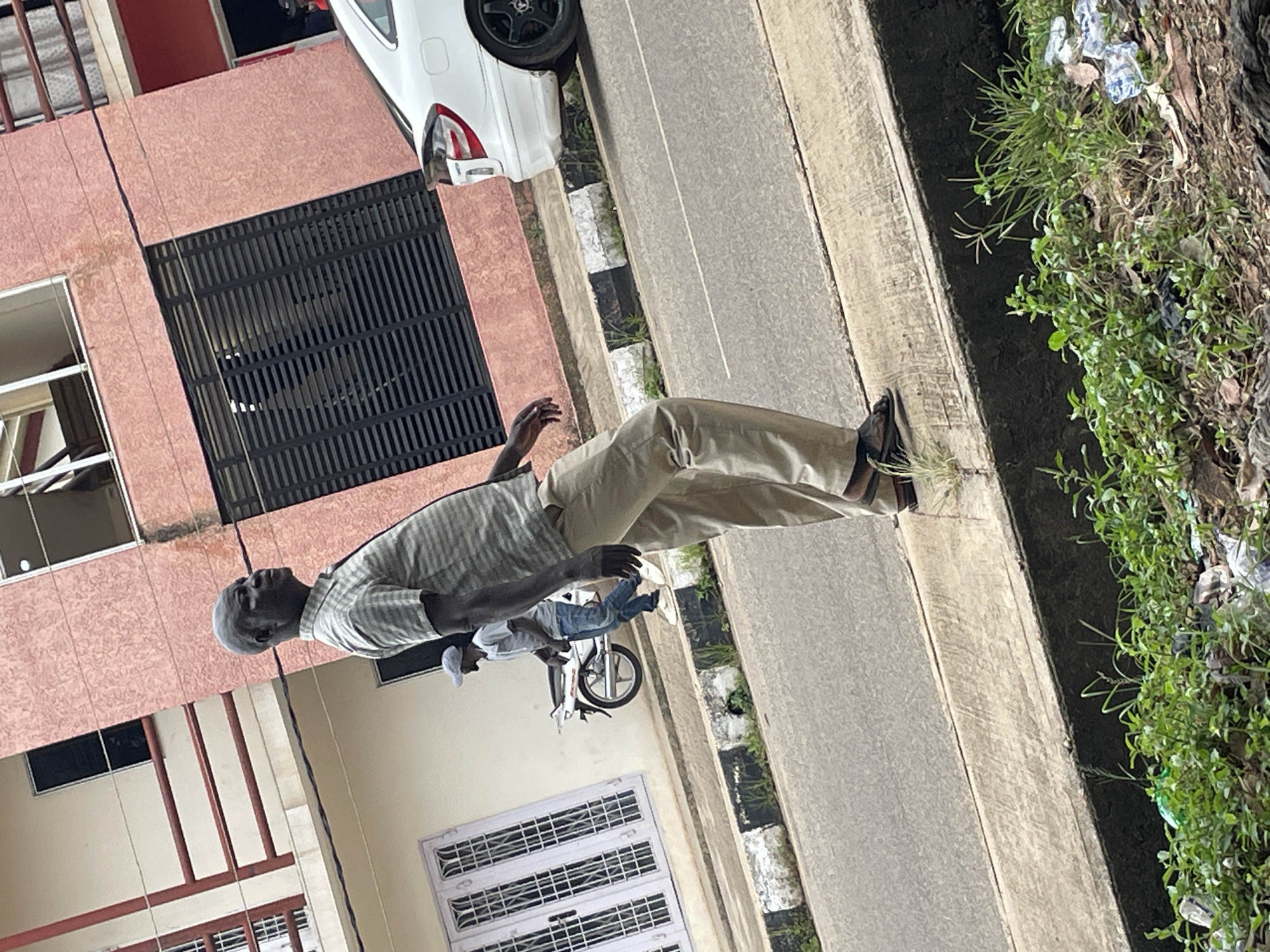 In Jos, Nigeria, a Black man in his late 40s or early 50s, with dark skin and a bald head, walks along the side of a street. He is wearing a light-colored, checkered short-sleeved shirt, khaki trousers, and dark sandals. His body is angled to the left, and he is looking forward. In the background, a white car is parked, and a motorcycle is visible with a person in a grey shirt and blue jeans standing next to it. The buildings in the background are painted in shades of pink and cream, with windows and balconies. The foreground shows a concrete curb separating the street from a patch of green grass and some scattered litter. The overall scene depicts a typical urban environment in Jos.