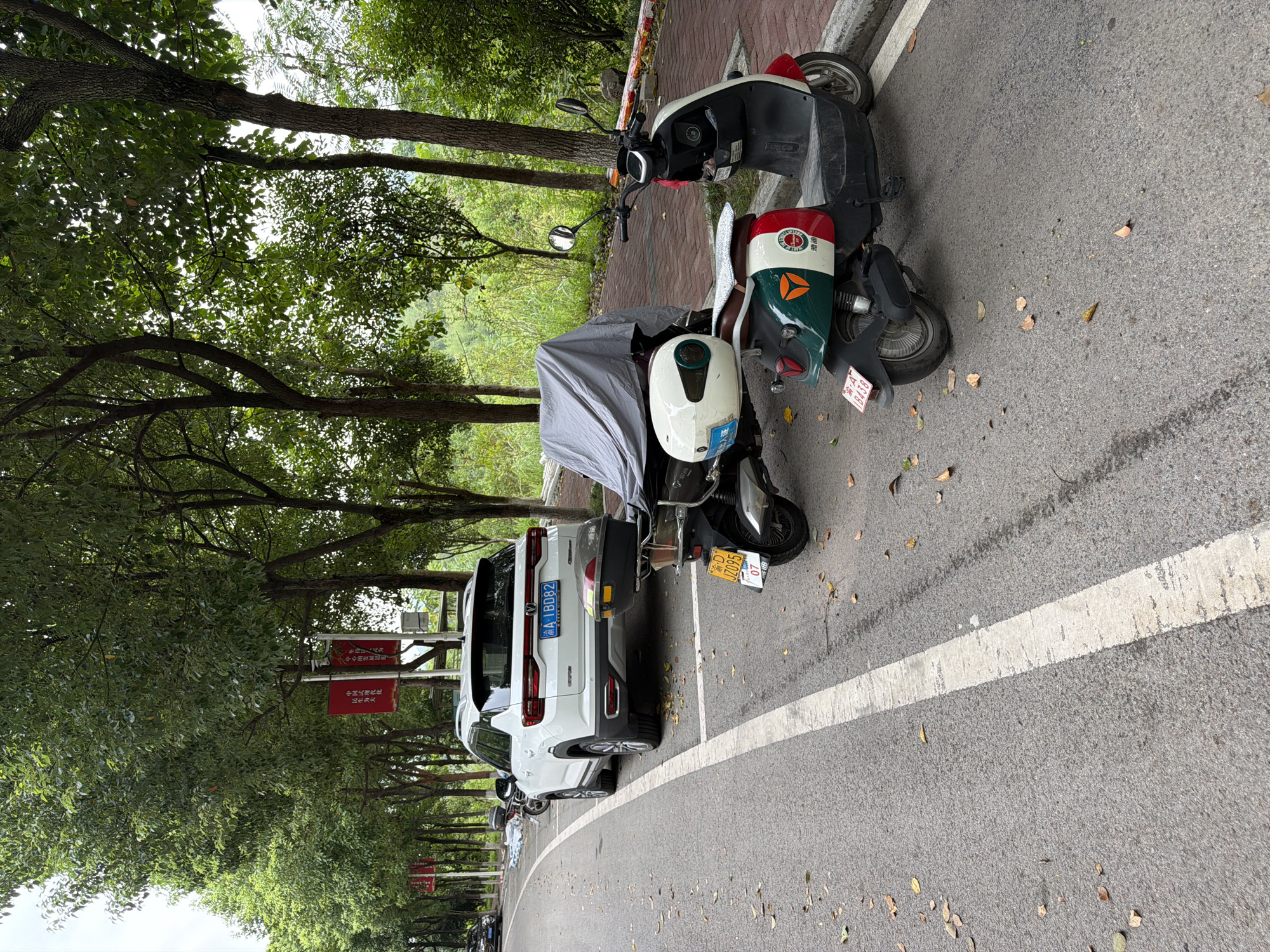 The image shows a street scene in the city of Jiangjin, China. In the foreground, two electric scooters are parked along the side of the road. The scooter on the right is red and white with green accents and a white storage box on the back. The scooter on the left is partially covered by a gray tarp. A white SUV is parked behind the scooters. The road is paved and has a white line marking the parking spaces. Trees line the street, and red banners hang from poles. The sky is overcast.