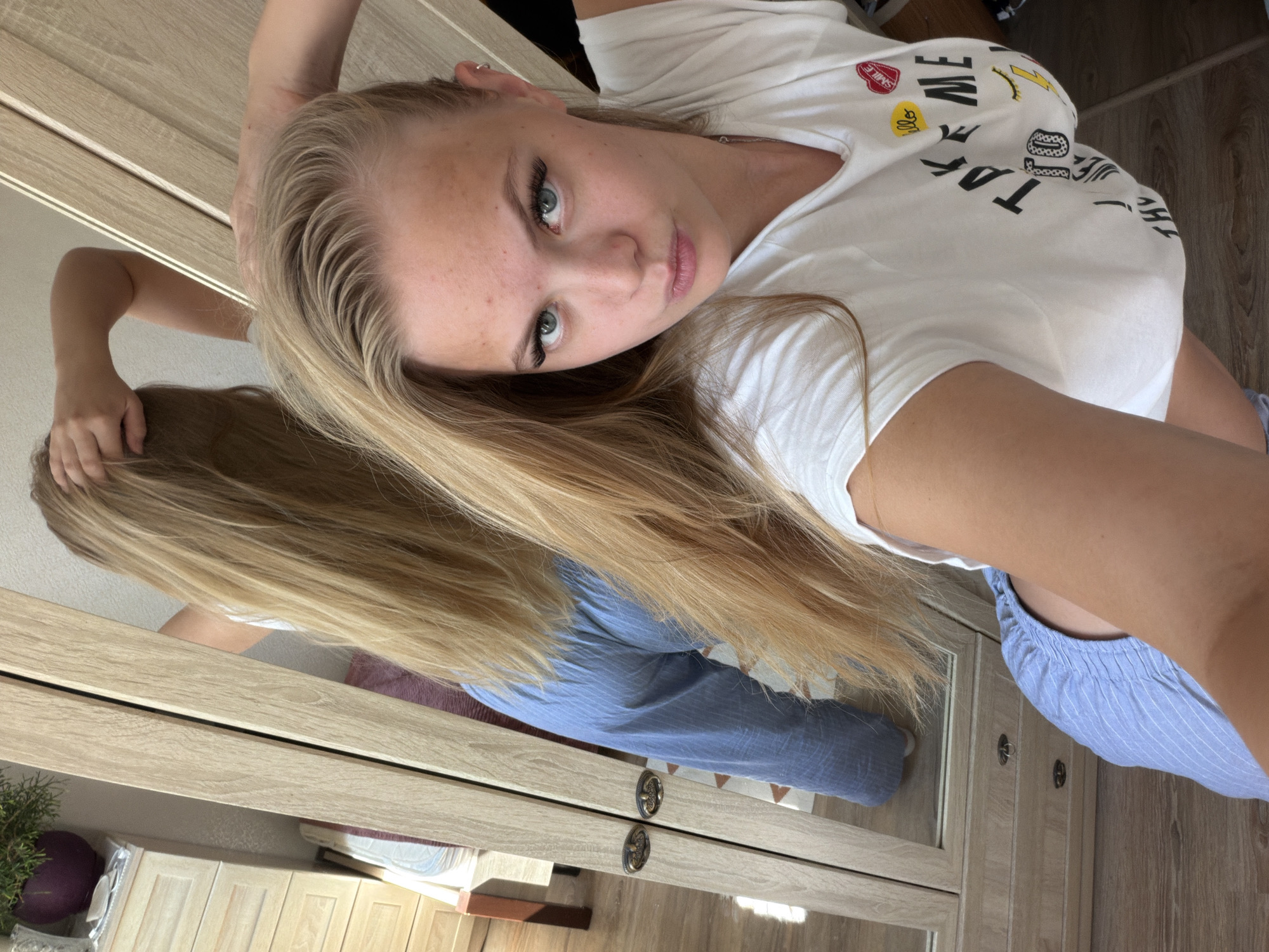 In a selfie taken indoors, a young woman with blonde hair and blue eyes is reflected in a mirror. She is wearing a white t-shirt with various cartoon-like images and text, including the words "ME," "SMILE," and "HELLO." Her hair is long and blonde, and it falls around her shoulders. She is looking directly at the camera with a relaxed expression. The background shows a wooden wardrobe with mirrors. The lighting suggests it is daytime. The woman appears to be in a bedroom or dressing area. The provided context indicates the location is Chaikovsky, Russia.