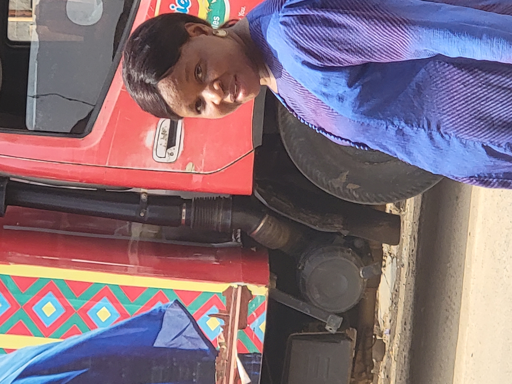 A woman in a blue striped shirt stands next to a red truck. The truck has colorful geometric patterns on its lower portion. The scene is set in the city of Maiburiji, Nigeria.