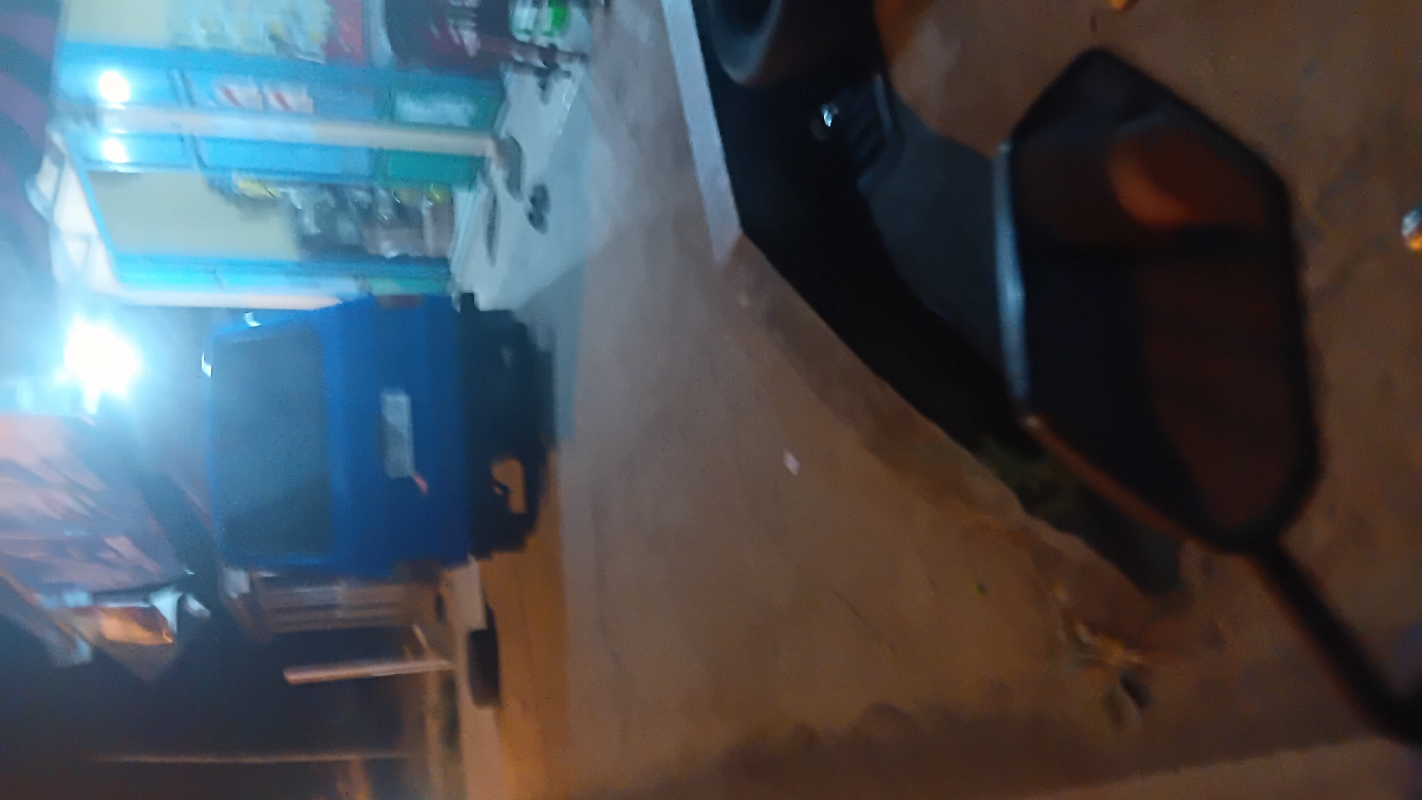 In Prabumulih, Indonesia, at night, a blue van is parked on a paved surface. The van's rear is visible, with a license plate and a thin orange object protruding from the right side of the license plate area. A black tire is visible on the ground to the left of the van. Behind the van, a building with a blue entranceway and yellow walls is illuminated by artificial light. In the foreground, a dark-colored object, possibly a motorcycle's rearview mirror, is partially visible. The overall scene is dimly lit, suggesting an outdoor urban environment.