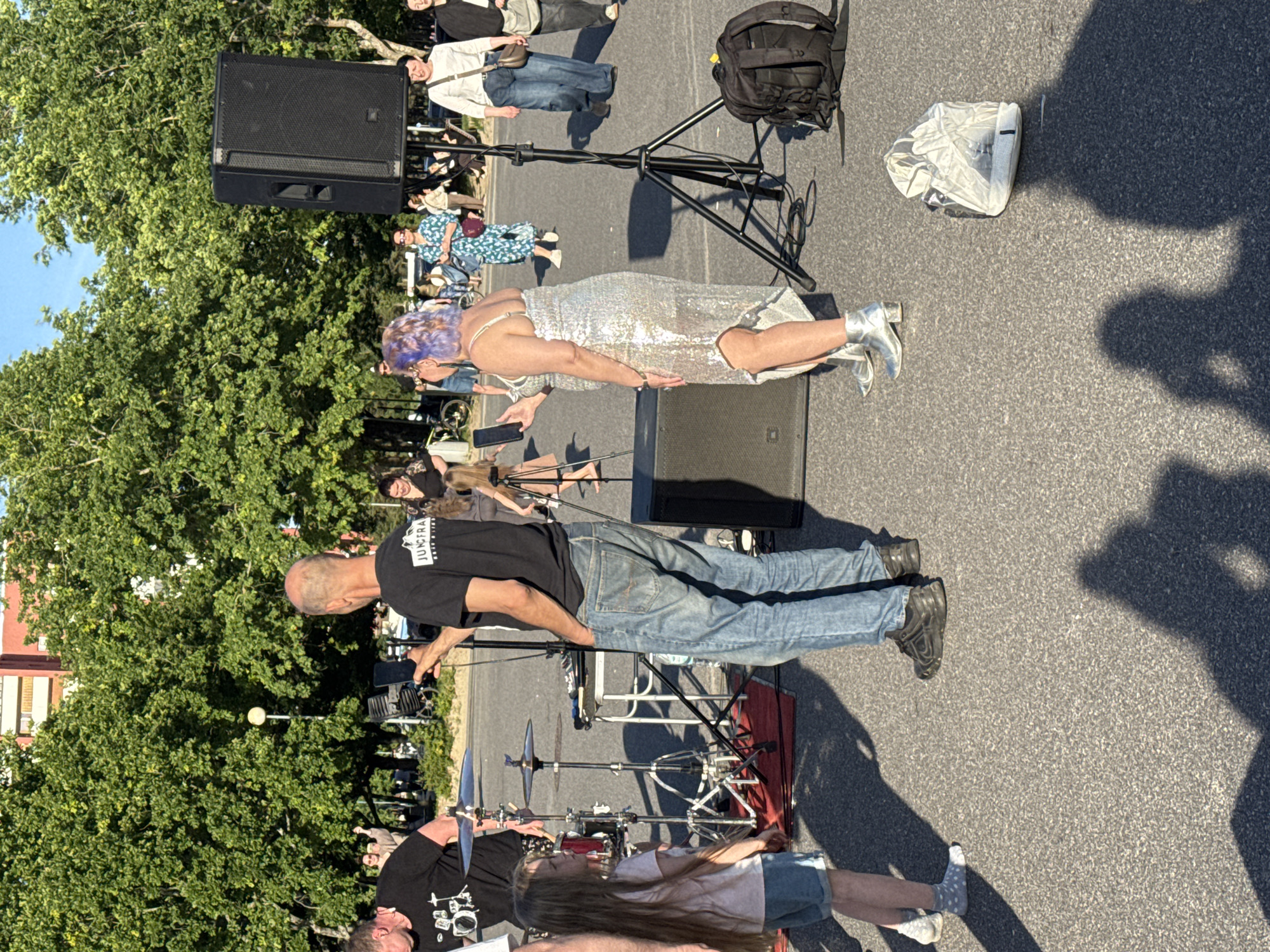 The image depicts an outdoor scene, likely a public event or gathering, with a group of people present. In the foreground, a young girl with long brown hair is looking to her left. Next to her, a man wearing a black t-shirt with a drum graphic and blue jeans is facing away from the camera, holding a mobile phone. To the right, a woman with purple hair is wearing a sparkly silver dress and silver boots. She is also holding a mobile phone and appears to be taking a picture or video.
Behind the people in the foreground, there is a drum set set up on a red mat, suggesting a musical performance is taking place or about to happen. A large black speaker is mounted on a stand to the right of the frame. Further in the background, more people are visible, some walking and others seated. Trees with green foliage dominate the background, with a building visible between the trees. The ground is paved with asphalt. The scene is bathed in sunlight, casting shadows.
While the image itself does not contain explicit clues about the location being Tallinn, Estonia, if this location information is provided contextually, it would indicate an outdoor event within the city. The overall impression is one of a casual, public gathering with a focus on music or entertainment.