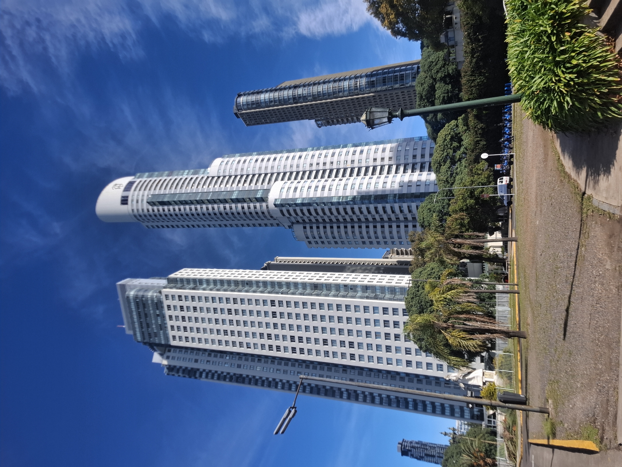 The image shows a row of tall, modern buildings in Buenos Aires, Argentina, viewed from street level. The tallest building is white and features a rounded top. The sky is a clear blue with wispy clouds. The foreground includes a sidewalk, a row of parked motorcycles, trees, and bushes. A lamppost is visible in the foreground, partially obscuring the buildings.