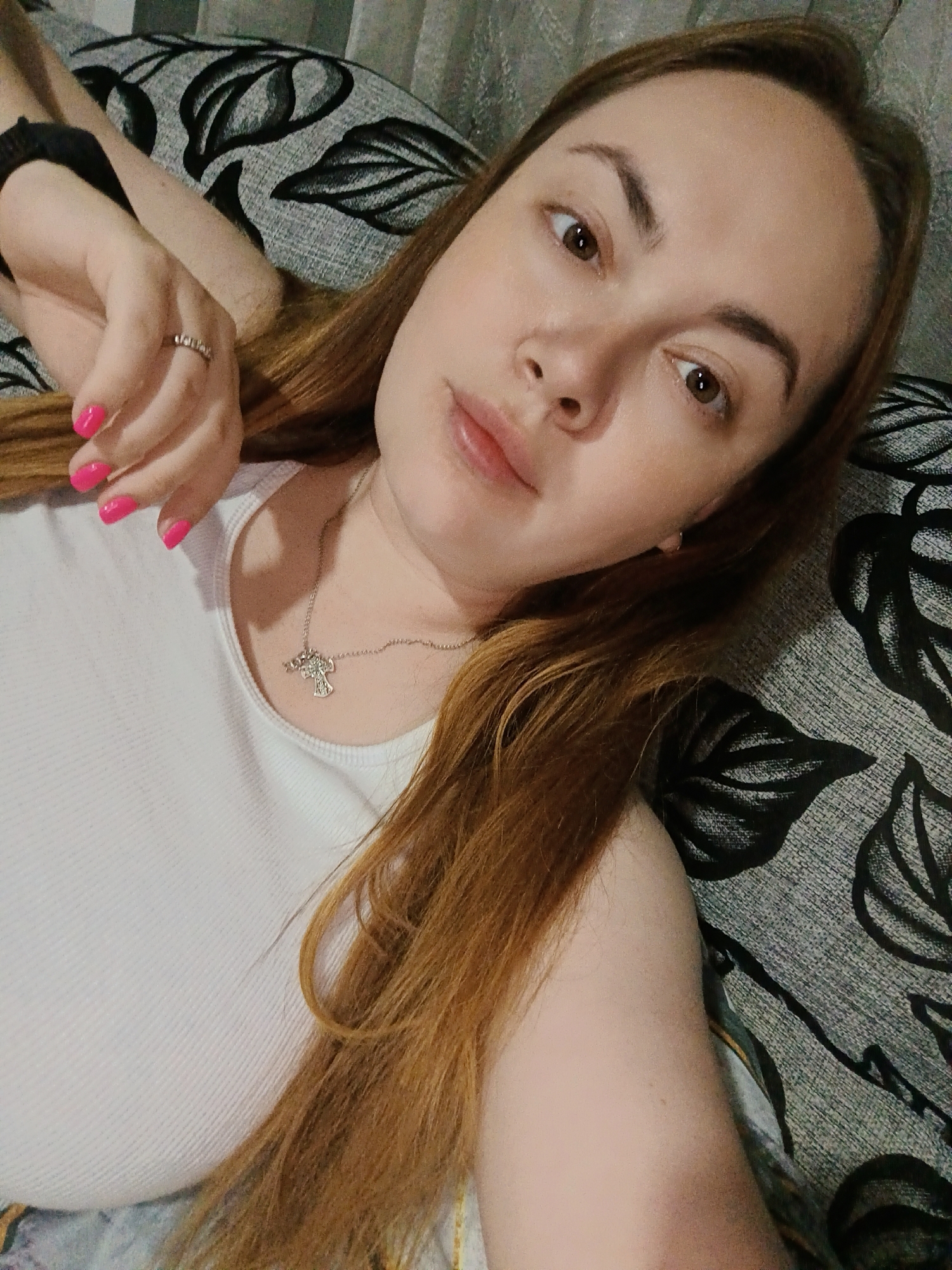 A young woman is depicted in a close-up, upward-angled selfie, lying down on what appears to be a sofa or bed with patterned cushions. Her head is tilted, and she looks directly into the camera with a neutral to slightly pensive expression.
The woman has long, straight brown or auburn hair that falls around her. She has fair skin, brown eyes, and well-defined eyebrows. Her lips are full. She is wearing a simple white ribbed tank top or camisole. Around her neck is a delicate silver chain necklace with a small, intricate silver pendant, possibly a stylized cross or angel. Her left hand is visible near her head, showing bright pink nail polish on her manicured nails and a silver ring on her ring finger. A black elastic hair tie or bracelet is on her left wrist.
The background consists of a dark gray fabric with a repeating pattern of stylized black leaves, likely a cushion or part of the furniture she is lying on. The lighting is soft and ambient, suggesting an indoor setting, possibly during the evening or in a dimly lit room. There is no visible text in the image. The scene takes place in Chaikovsky, Russia.