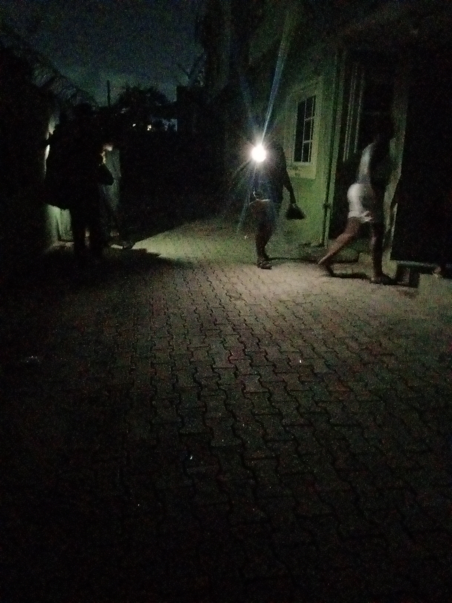 In the image, several people are walking in a dimly lit alley in Ado, Nigeria. The alley is paved with interlocking bricks. One person in the center is wearing a headlamp, illuminating their path. A building with a window is visible on the right side of the alley. Other people are also present, but their features are obscured by the darkness.