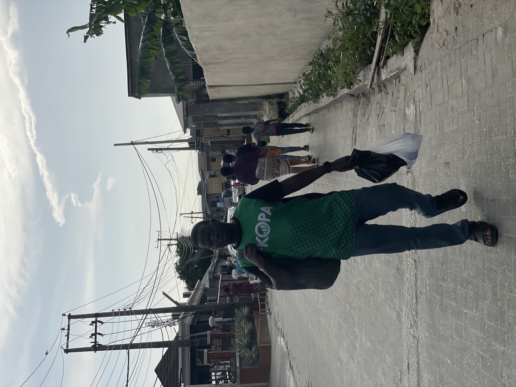 A man stands on a paved street in Iranla, Nigeria, wearing a green t-shirt with the word "KOPA" printed on it. He holds a black plastic bag in his right hand and a larger bag in his left hand. In the background, several people walk down the street. Buildings line the street, and power lines are visible overhead.