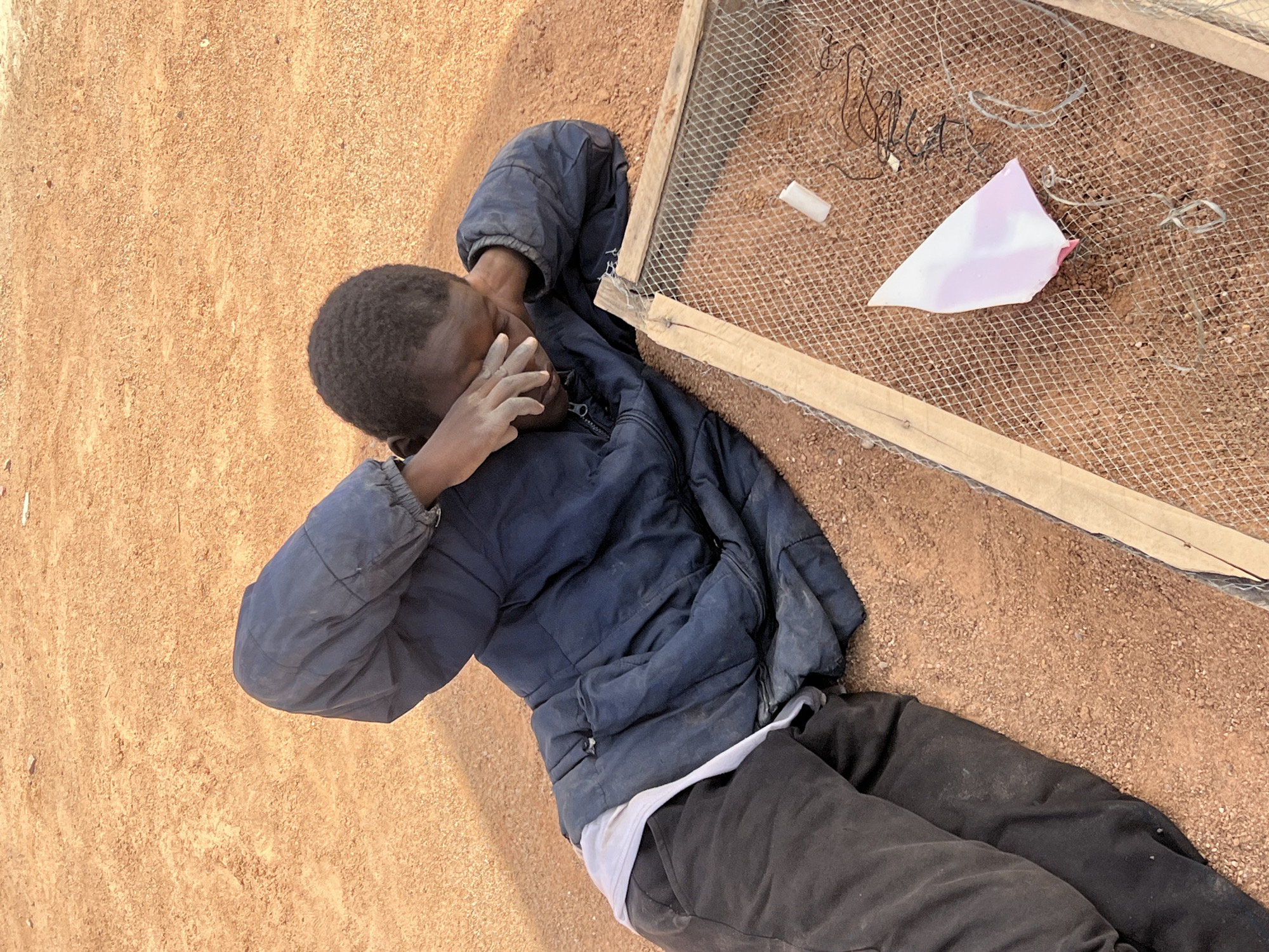 The image shows a person lying on a reddish-brown, granular ground surface, possibly sand or dirt. The person is wearing dark trousers, a white undershirt and a dark blue padded jacket, and is turned slightly away from the camera, with their head near a rectangular wooden frame with wire mesh. The person's hand is near their face, obscuring the view. Several small white objects, a torn piece of pink paper, and lengths of black wire are resting on the wire mesh within the frame. The location is assumed to be Barakin Lerawi, Nigeria, based on the prompt.
