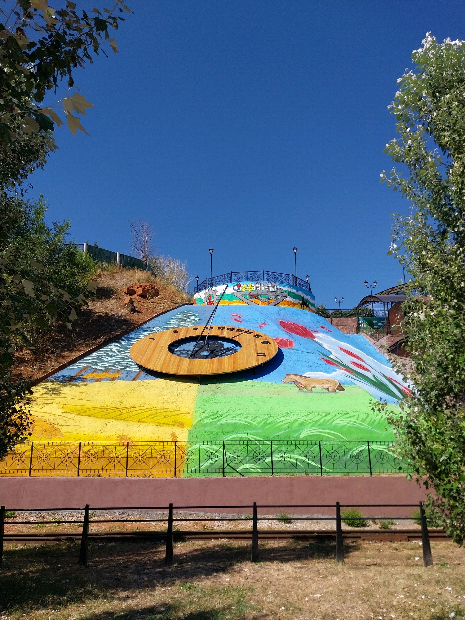 A vibrant mural covers a hillside in Orenburg, Russia, depicting various natural elements and a large sundial. The mural is divided into sections with distinct colors and imagery. The bottom section features a yellow landscape with depictions of wheat or grass, transitioning to a bright green grassy area. Above this, a blue section shows wavy lines representing water, and further up, a larger blue area with red and white abstract shapes that could be flowers or leaves. A prominent feature is a large, wooden sundial with black hour markers and hands, positioned in the middle of the blue watery section. A light brown horse is painted in the green section. At the top of the hill, a fence with ornate black railings encloses a walkway or observation area, and streetlights are visible. Trees with green foliage frame the scene from the left and right. The sky is a clear, deep blue. The ground in front of the mural is covered with dry grass and is bordered by two rows of dark-colored fences. Railway tracks are visible between the fences.
