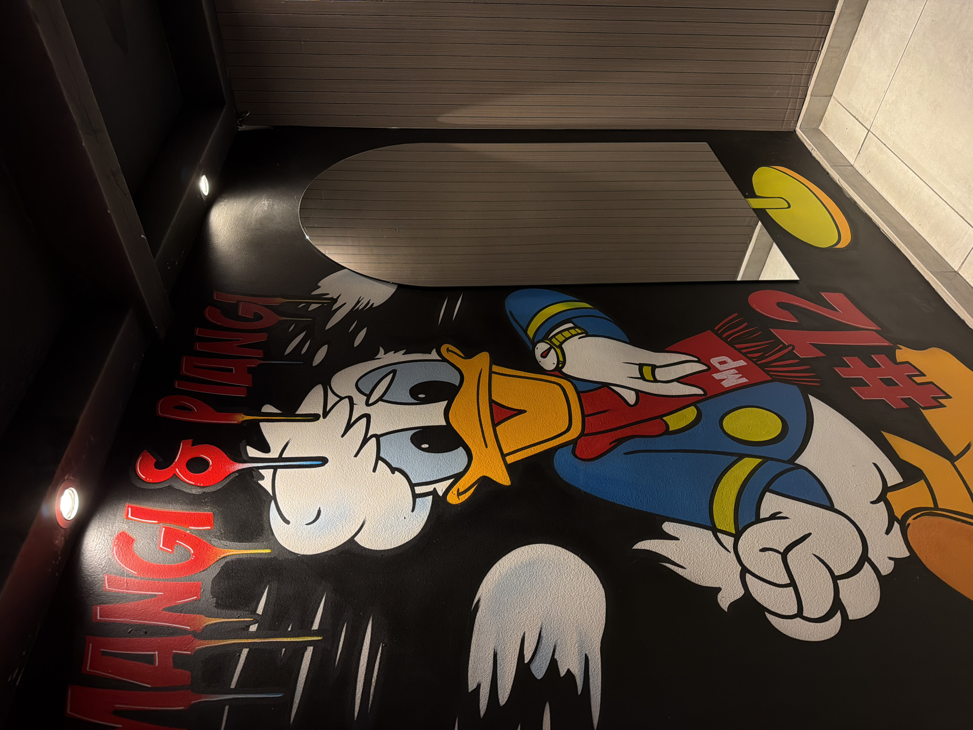 An angled photograph captures a wall mural of Donald Duck on a black background. Donald is wearing a blue jacket with yellow buttons and a blue hat with yellow stripes. He is holding a playing card in his hand. The words "MANGIA & PIANGI" are vertically oriented on the left. The number "72#" is vertically oriented on the right. Above the mural, there is a narrow section of horizontally-striped wall. The photo was taken inside, in Varna, Bulgaria.