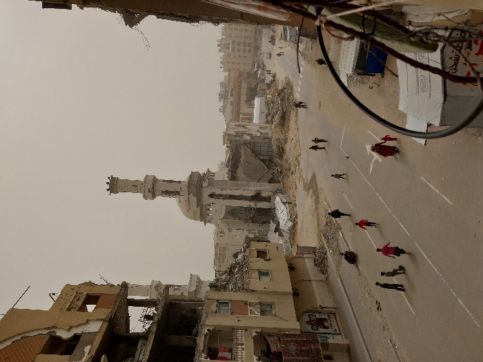 The image displays a severely damaged urban street scene in Khan Yunis, Palestinian Territory, viewed from an elevated vantage point under an overcast or dusty sky. Dominating the background is a large mosque, featuring a tall, slender minaret and a dome, both extensively damaged with visible structural collapse and accumulated debris at their base. Buildings flanking the street are significantly ruined, exhibiting crumbling facades, exposed interiors, and missing sections. On the left, a multi-story residential-style building shows partial destruction, with personal items visible on balconies. The two-lane asphalt road has faded white lane markings and a directional arrow. Approximately 15 individuals, including adults and children, are present on the street: a woman in a red garment walks with a small child, a group of children appear to be playing, and other individuals walk or stand. The areas adjacent to the road are covered in rubble, dirt, and debris from destroyed structures. In the immediate foreground right, a metal railing partially obstructs the view, with a white tent-like structure featuring Arabic text and a smaller blue-and-white kiosk located below it. Distant vehicles are faintly visible on a far street.