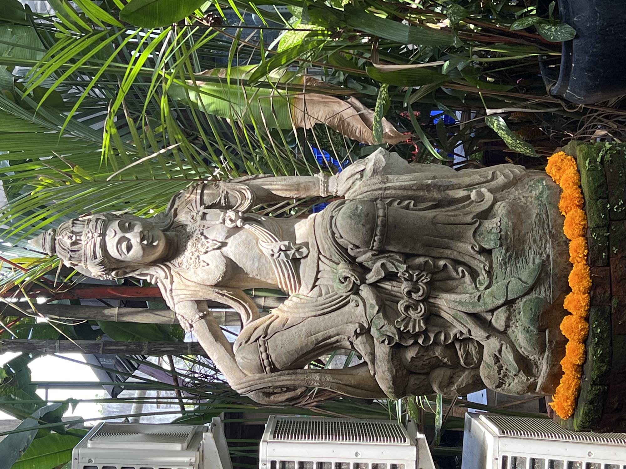 The image depicts a weathered stone statue of a Balinese deity, adorned with a string of bright orange marigolds, resting against a mossy stone wall or platform. The statue is surrounded by dense tropical foliage, including palm fronds and other leafy plants. Below the statue are several white air conditioning units. The location is Peliatan, Indonesia.