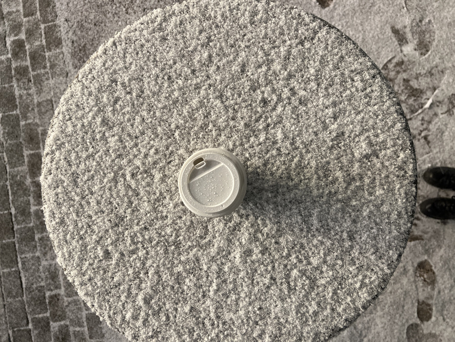 An overhead forensic view captures an outdoor winter scene in Saint Petersburg, Russia. A circular table, approximately 0.75 meters in diameter, is covered with a uniform, thin layer of granular snow. Centered on the snow-dusted table is a light gray, possibly reusable, coffee cup with a closed flip-top lid, exhibiting small water droplets on its surface. Beyond the table, the ground is similarly snow-covered, revealing the texture of cobblestone pavement. In the lower portion of the frame, the tips of two dark-colored shoes are visible, positioned on the snow-covered ground, alongside several partial footprints impressed into the snow. No other individuals are present.