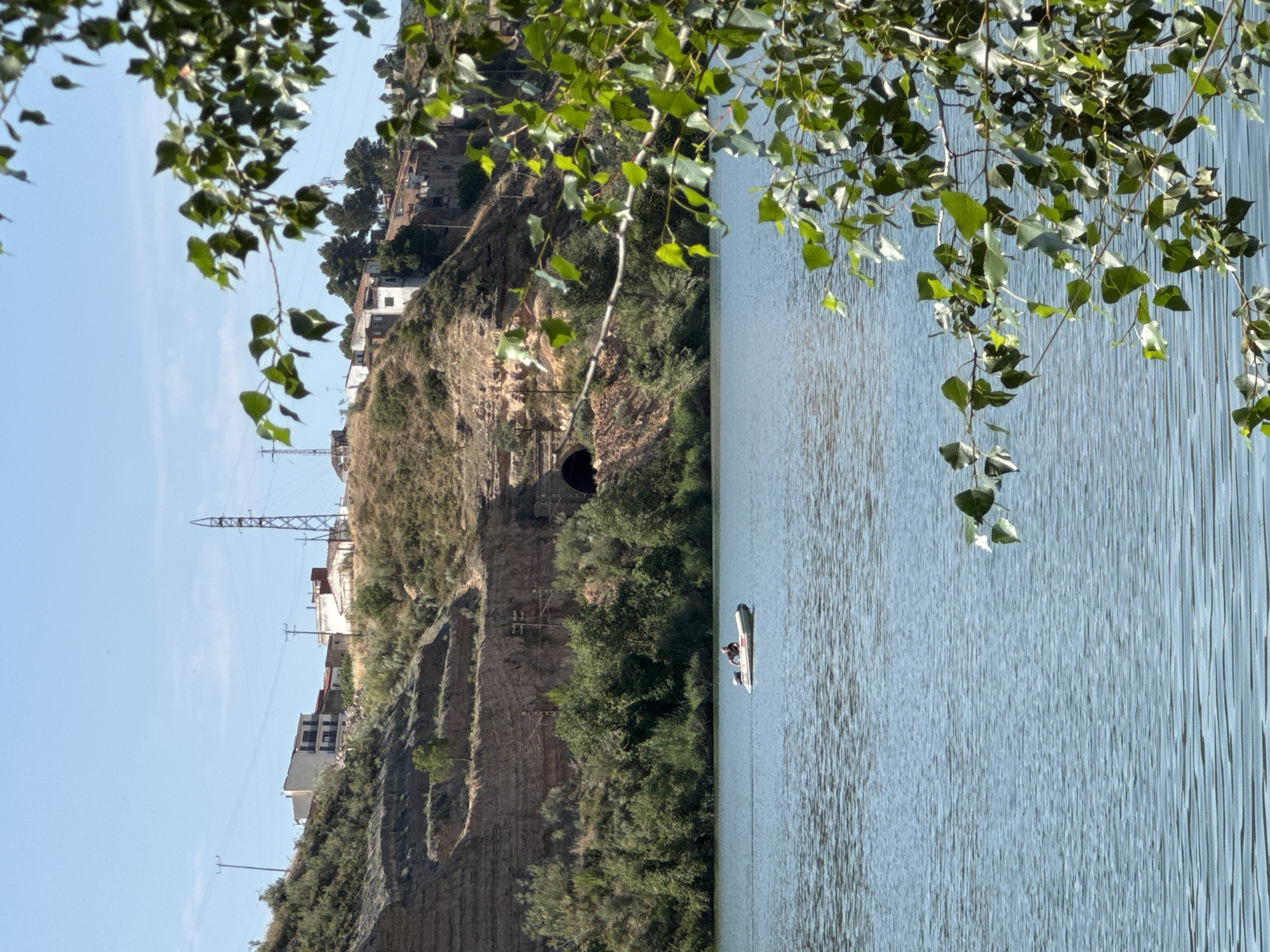 A wide shot captures a serene, blue body of water under a clear sky. In the distance, a brown, layered hillside rises, dotted with green foliage and a few buildings. A dark, arched tunnel is visible carved into the hillside. A small, white inflatable boat with an outboard motor is positioned in the middle of the water. A single person, wearing dark clothing, is seated in the boat, facing towards the left. The foreground is partially obscured by the lush, green leaves of a tree, creating a natural frame for the scene.