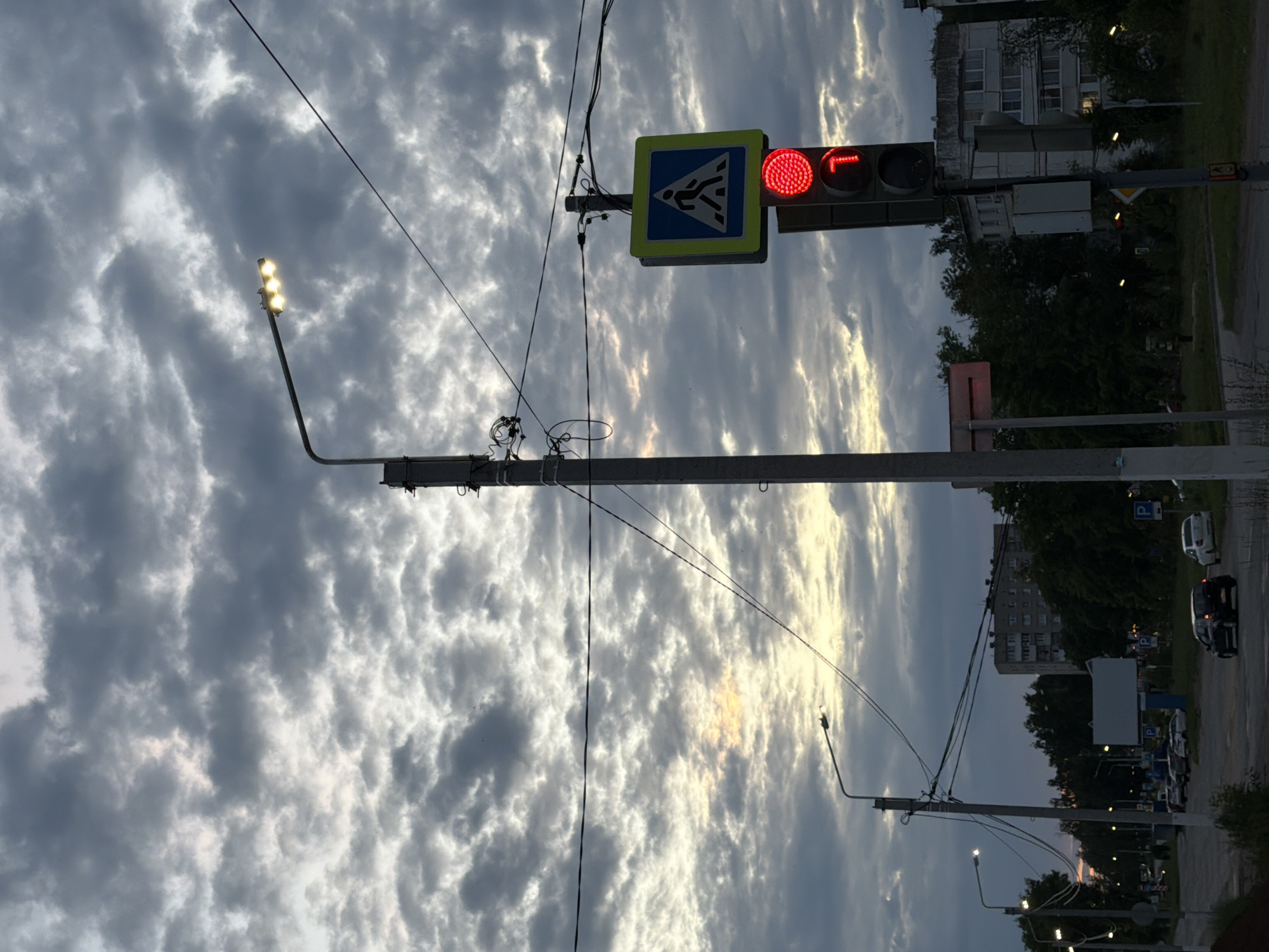 The image is a medium shot of a city street at dusk. The sky is filled with dramatic, textured clouds, with a hint of yellow light peeking through the overcast. A tall, grey utility pole dominates the foreground, supporting a street lamp with three illuminated bulbs and a network of electrical wires. To the right of the utility pole, another pole holds a traffic light, which is currently red, and a blue and yellow pedestrian crossing sign.
In the background, the street curves to the left, lined with trees and buildings. Several cars are visible on the street, parked and in motion. The overall atmosphere is somber and transitional, as day gives way to night.