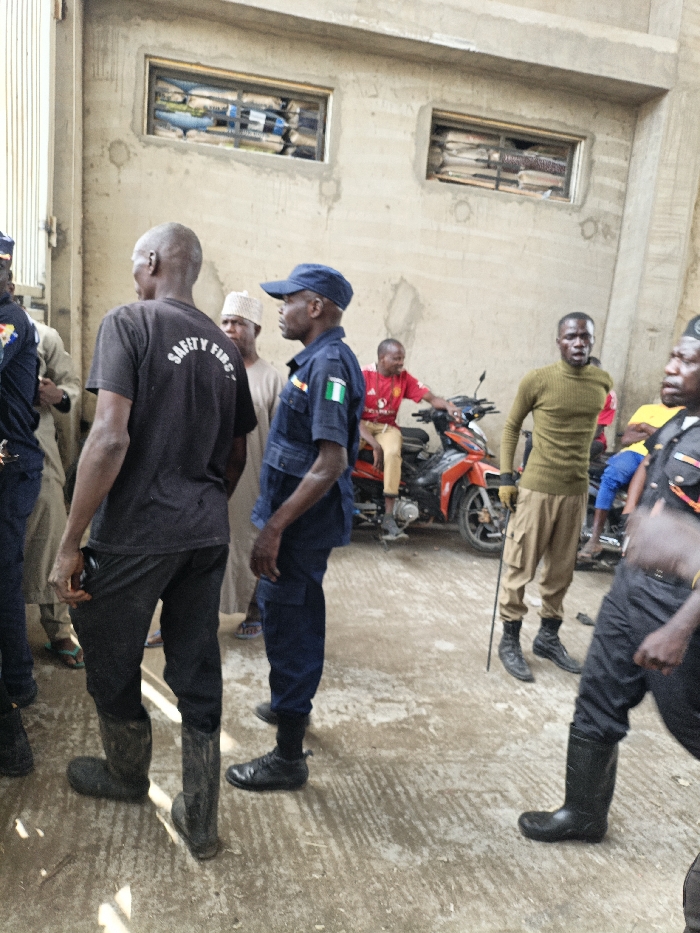 The image depicts an outdoor scene in Fagge C, Nigeria, showing approximately eight adult males on a concrete surface against a light-colored, possibly unfinished concrete building. The building features two rectangular window openings, each containing horizontally stacked bags, possibly sacks of grain or cement.
In the foreground, a male with his back to the camera wears a black t-shirt with "SAFETY FIRST" printed in white, black trousers, and tall black rubber boots. To his right, a male in a dark blue uniform, including a cap and a Nigerian flag patch on his sleeve, stands facing right. Behind these two, a male wearing a red shirt is seated on an orange and black motorcycle. To the right of the uniformed man, another male stands upright, dressed in an olive green long-sleeved turtleneck shirt, light brown cargo pants, and dark lace-up boots, holding a long, slender dark stick with its tip on the ground. A male in a partial dark uniform with yellow elements and black rubber boots is visible on the far right. Another individual wearing a light-colored traditional cap is visible in the middle ground. The individuals are standing or seated, appearing to be gathered or observing.