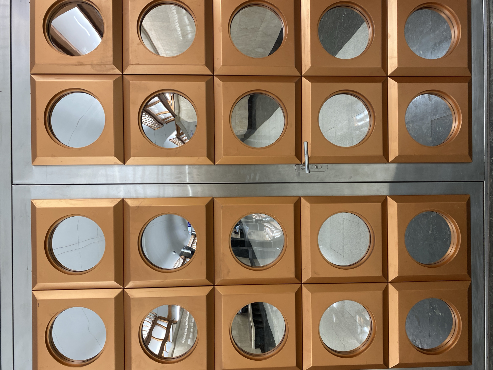 The image displays a decorative wall or door feature consisting of a grid of twenty square, bronze-colored panels, arranged in four rows and five columns. Each square panel is inset with a circular mirror. The entire assembly is bordered by a brushed silver-metallic frame on all four sides and divided horizontally in the middle by a similar metallic band.
No direct activity is taking place; the image captures a static architectural or design element. The various circular mirrors reflect different perspectives of an unseen interior space. Reflections show a mix of architectural features, including ceilings, walls, and what appears to be patterned flooring. Some mirrors clearly reflect wooden chairs or other furniture, while others capture ambient light and shadows, indicating an indoor setting.
Notable details include the warm, uniform bronze color of the square panels, which possess a slightly textured, matte finish. The metallic framing and central band have a subtle sheen. On the right side of the central metallic band, there is a small latch or hook mechanism. While the location context of Saint Petersburg, Russia, is provided, there are no specific visual cues within the image itself to confirm this. The lighting is consistent with an indoor environment, but the time of day is not discernible. No visible text can be clearly identified; there are some faint, illegible markings on the metallic band near the latch.