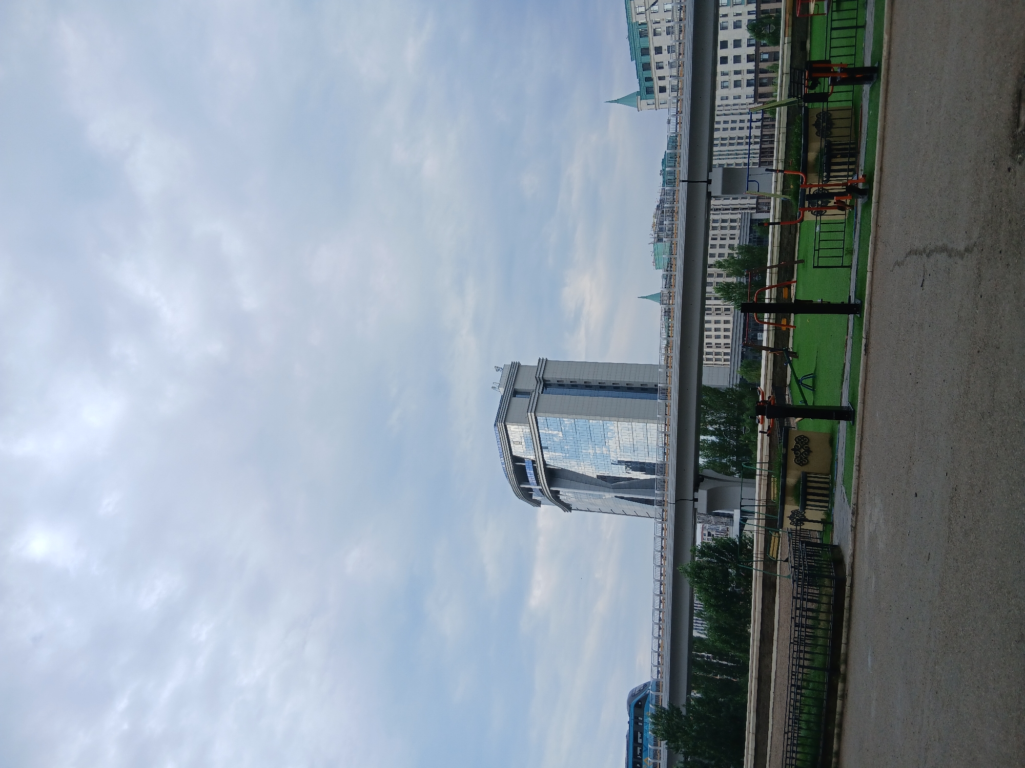 This image depicts a scene in Astana, Kazakhstan, focusing on a modern cityscape with a prominent elevated metro line. In the foreground, a paved road transitions into a fenced-off area containing a children's playground with swings and other equipment, situated on a green surface. Behind the playground, a concrete metro line structure runs horizontally across the frame. A blue and silver train is visible on the metro line, moving from left to right.
Dominating the middle ground is a distinctive, modern skyscraper with a curved, glass-paneled facade that reflects the overcast sky. To the right of this skyscraper, other multi-story buildings with European-inspired architectural elements, including green roofs and pointed turrets, are visible. Dense green trees line the area beneath the metro line, creating a contrast with the urban structures. The sky is filled with heavy, grey clouds, suggesting overcast weather. The overall impression is one of contemporary urban development integrated with recreational spaces.