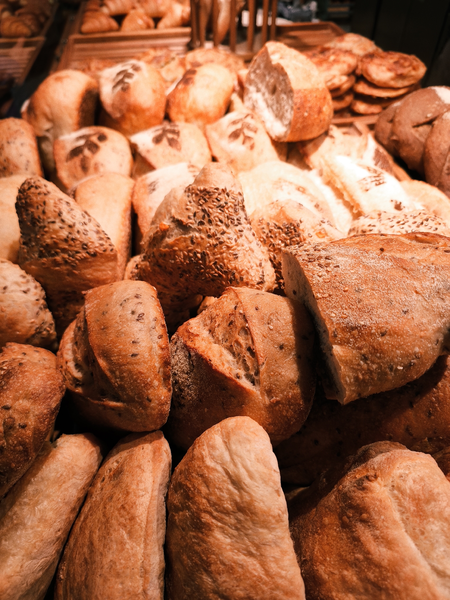 A close-up, eye-level, medium shot captures a generous display of freshly baked bread, likely at a bakery in Svetlogorsk, Russia. The bread is arranged in a dense, abundant pile, with various shapes and sizes filling the frame. There are several round loaves with visible scoring on their crusts, some of which are generously sprinkled with seeds, possibly sesame or flax. Other loaves appear to be more elongated, baguette-style, also with seed toppings and a rustic, textured surface. The crusts exhibit a rich, golden-brown hue, suggesting they have been recently baked. The lighting is warm and inviting, highlighting the inviting texture and color of the bread. The background is largely out of focus, but hints at more bread or bakery items, implying a retail setting. No people or animals are visible in the frame.